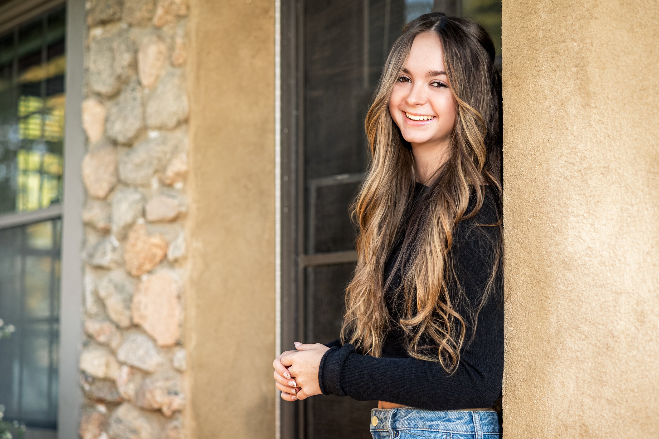 A young woman with long wavy hair smiling and leaning against a yellow stucco wall outside a building.