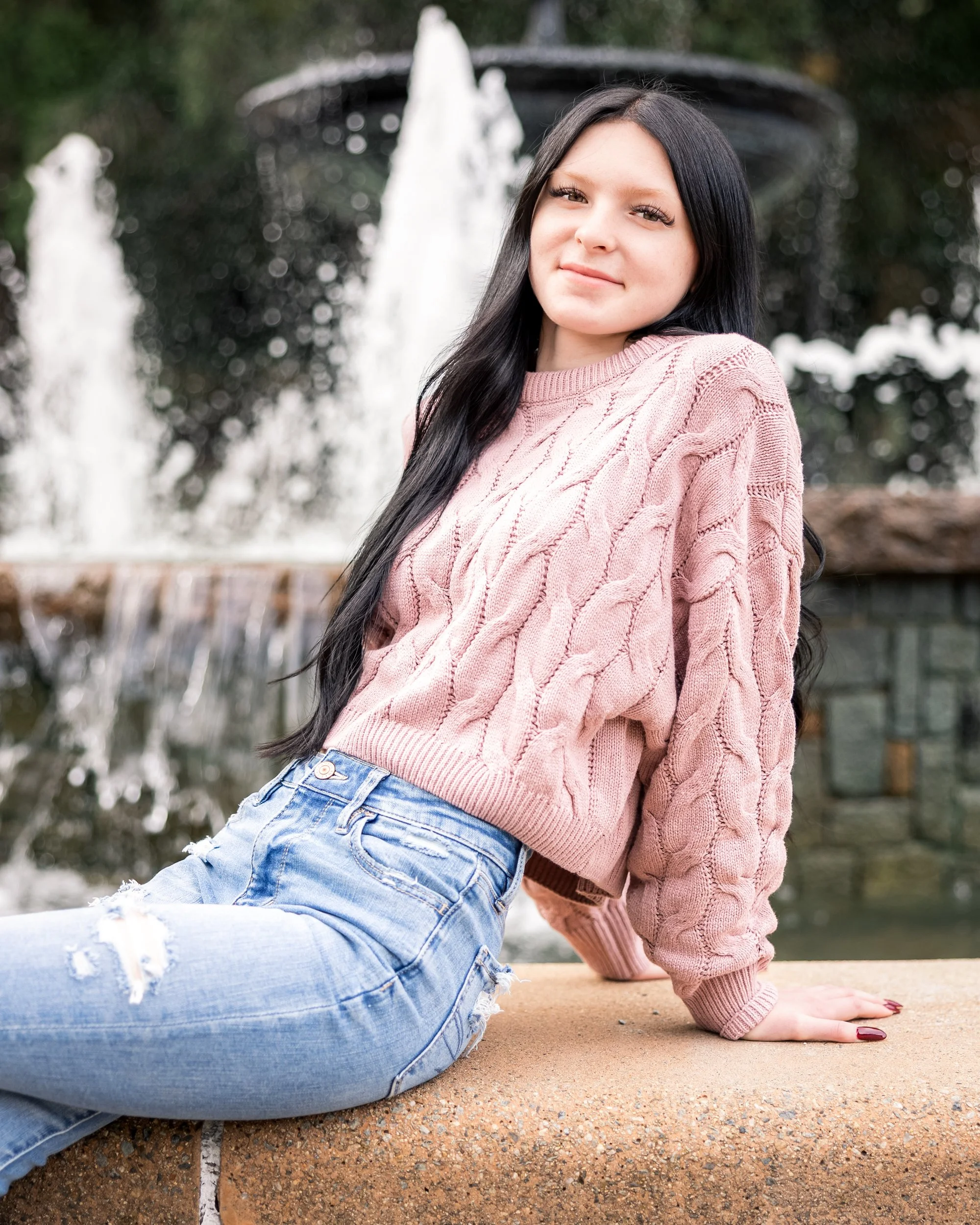A young woman with long black hair sitting on a stone ledge in front of a water fountain, wearing a pink cable-knit sweater and ripped light blue jeans.
