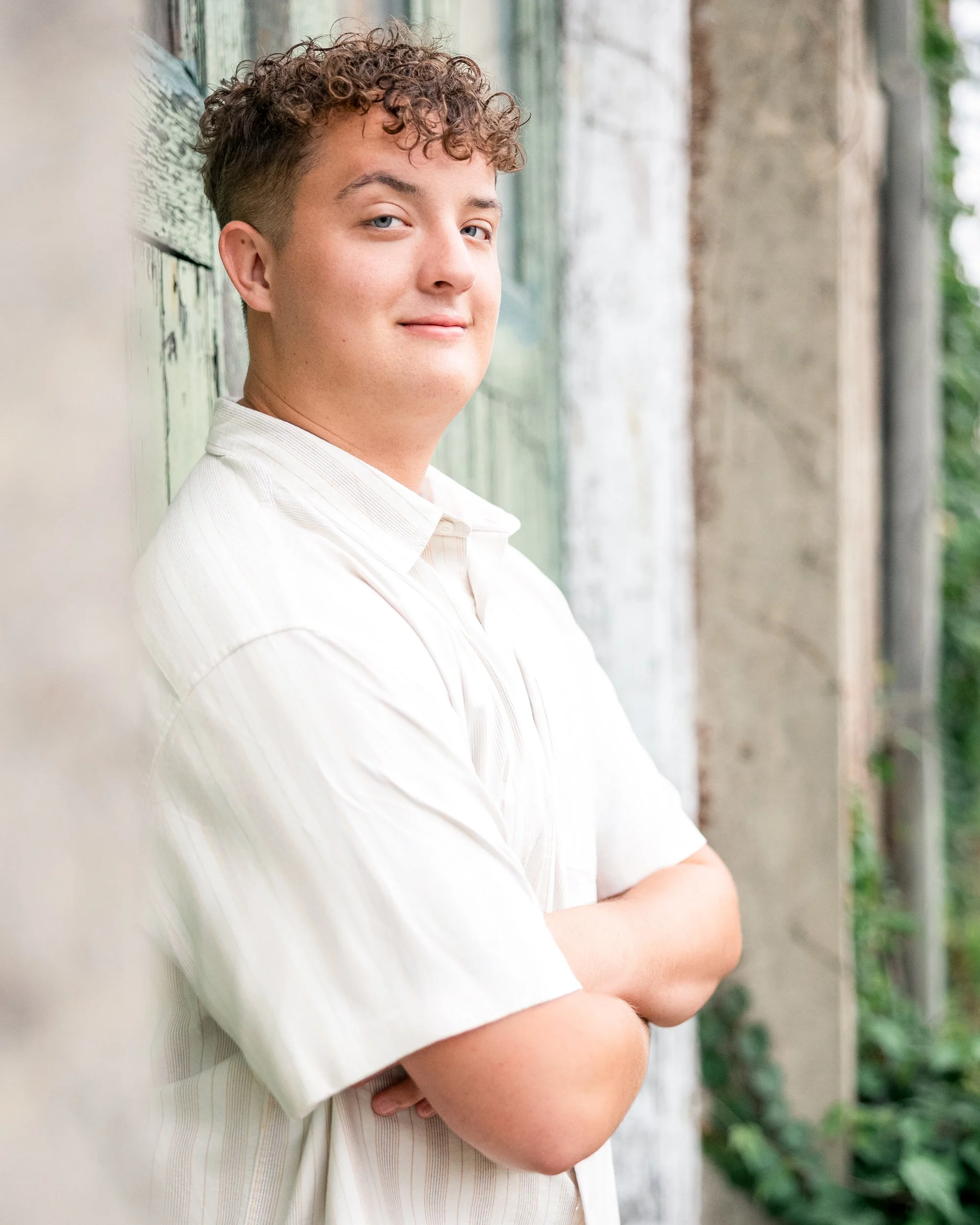 A young man with curly hair wearing a light-colored shirt, standing outside with arms crossed, leaning against a wall with a blurred background.