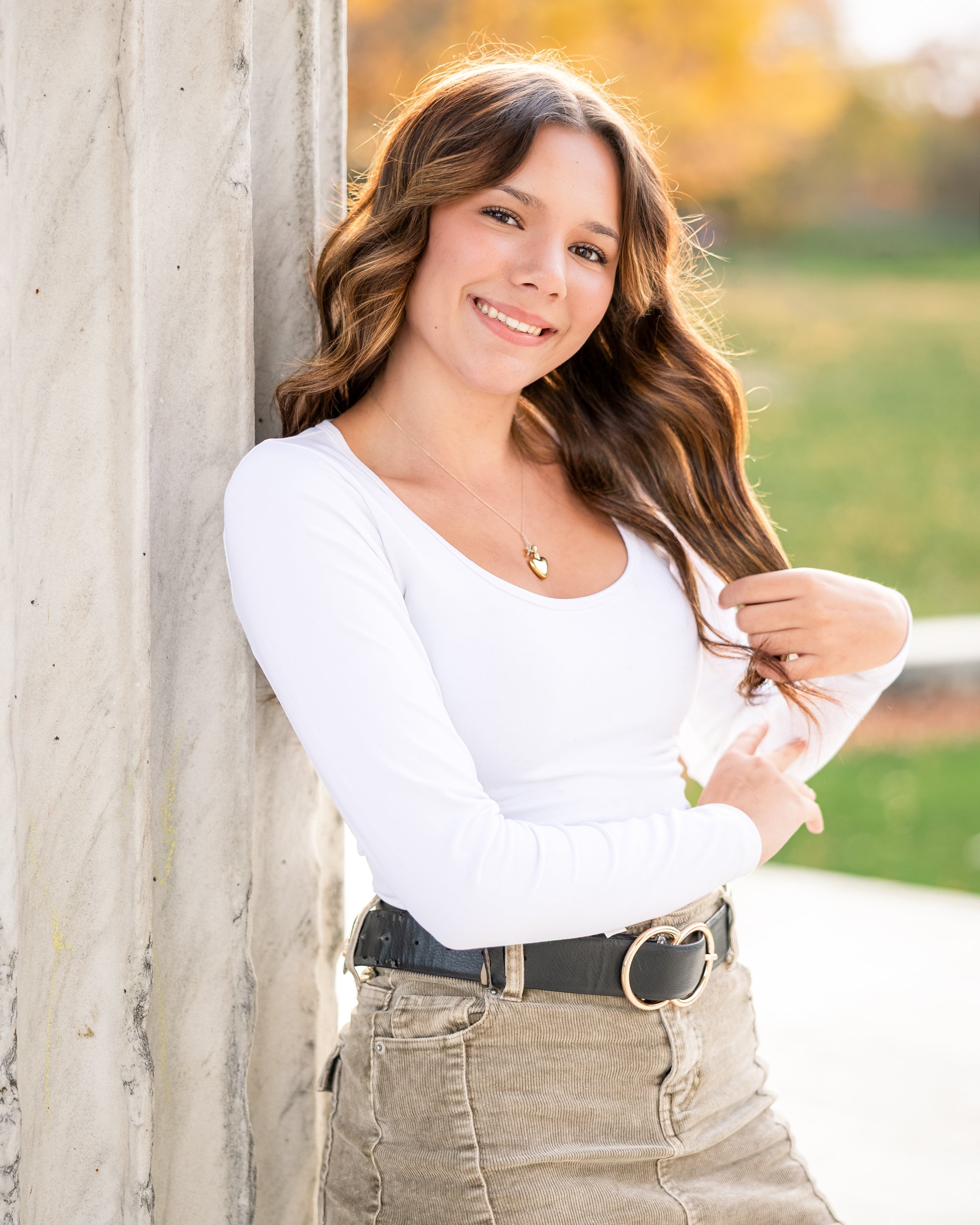 A young woman leaning against a concrete wall outdoors, smiling, with autumn foliage in the background.