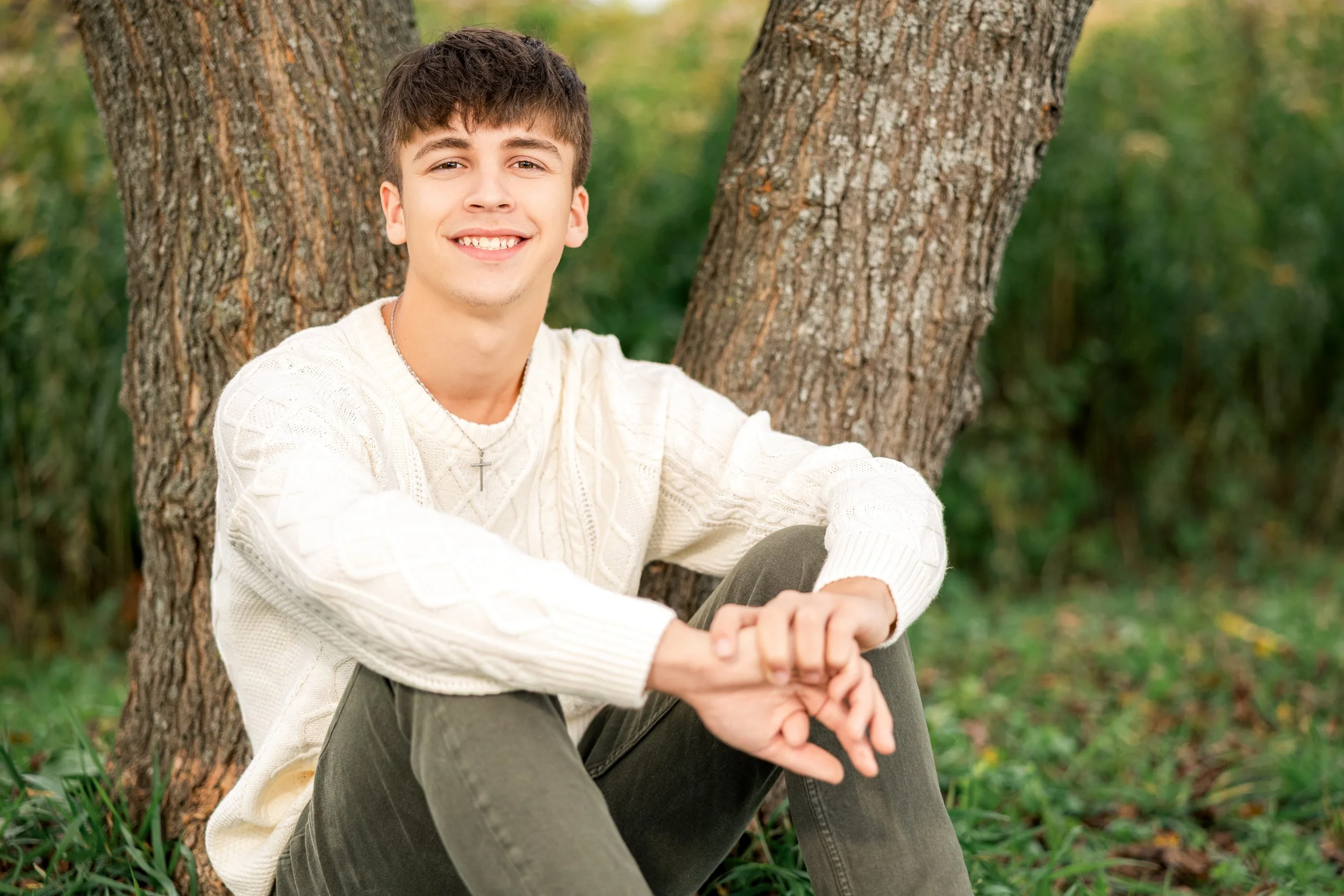 Smiling young man sitting outdoors near a tree with green foliage, wearing a cream cable-knit sweater, dark pants, and a cross necklace.