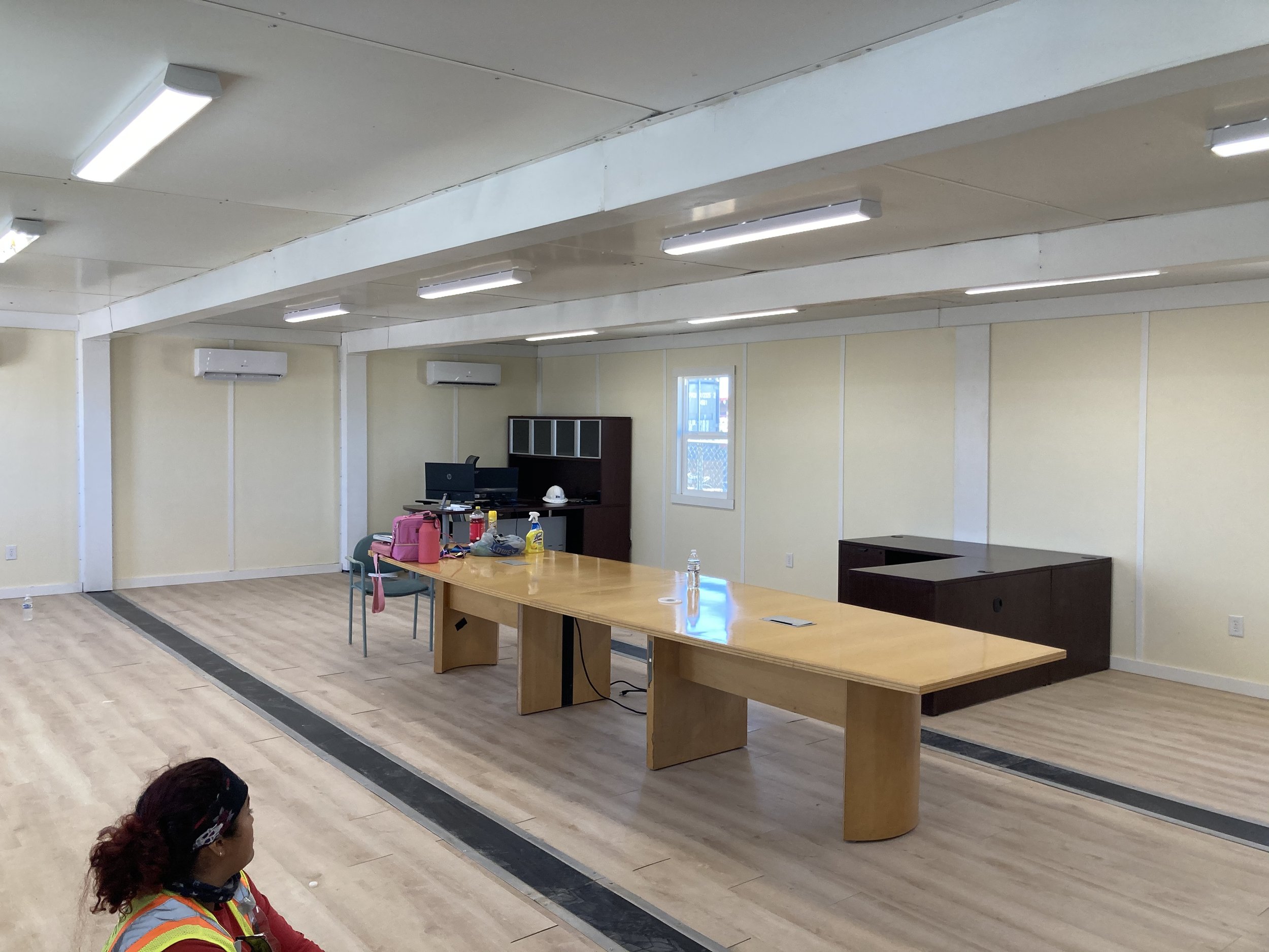Empty office space with a large wooden conference table, two chairs, a desk with two computer monitors, and miscellaneous items. Light-colored walls, ceiling with fluorescent lights, window, and wall-mounted air conditioners.
