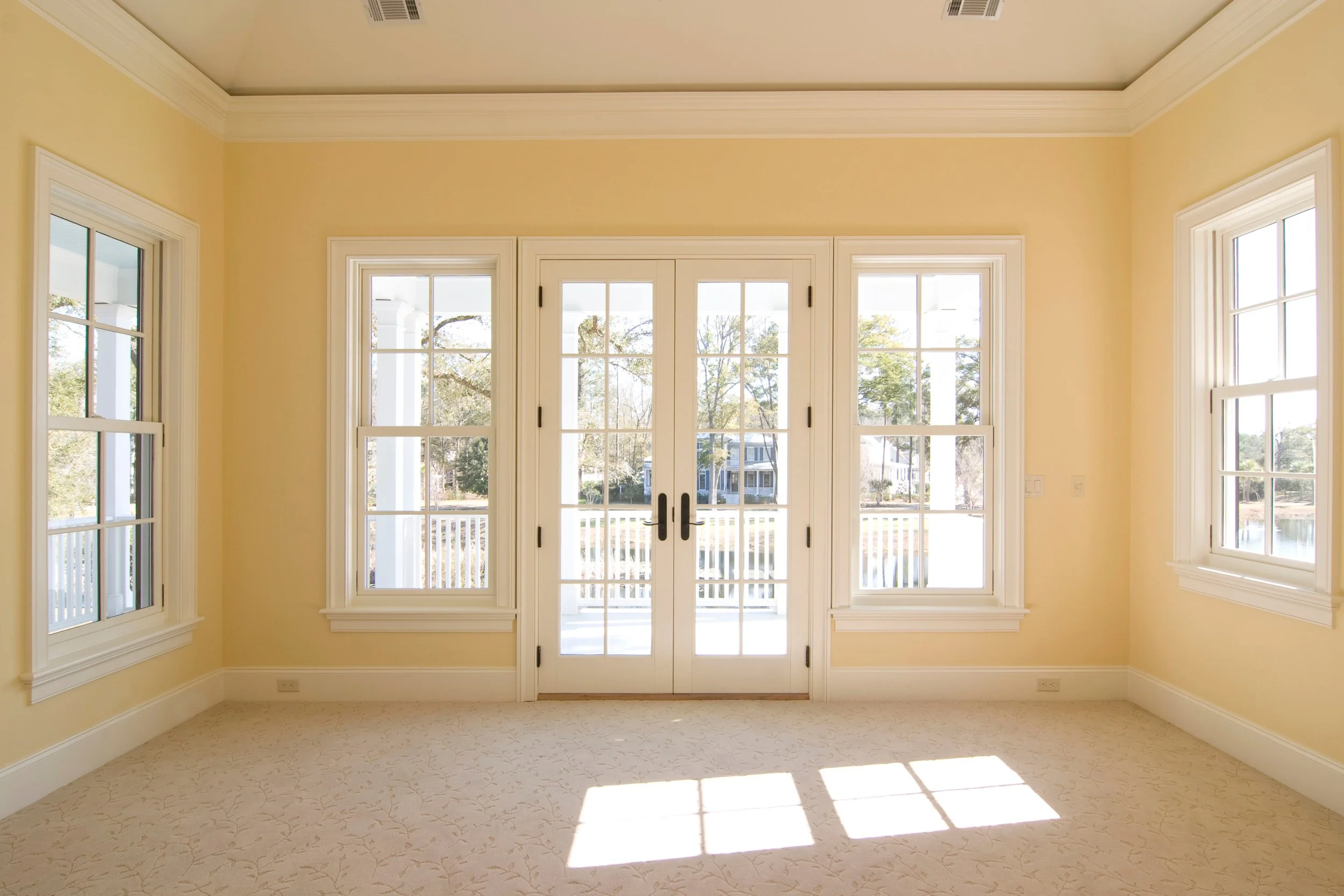 Bright empty room with 4 large white-framed windows and a double glass door leading to an outdoor porch, yellow walls, white crown molding, and a beige patterned carpet.