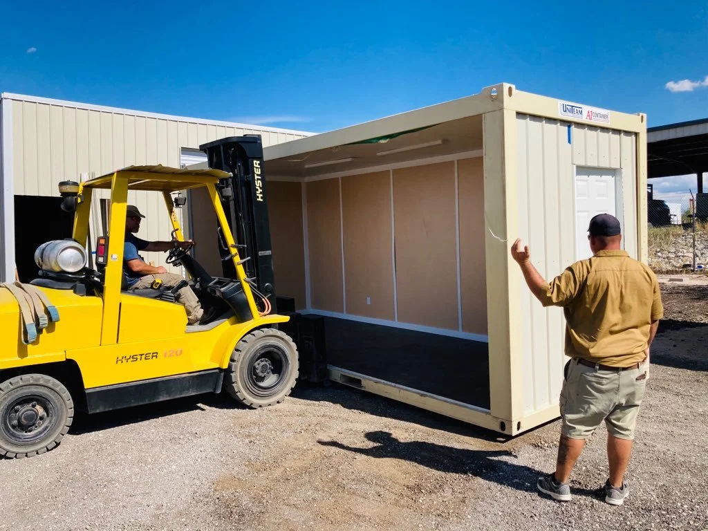A man guiding a forklift operator as they position a beige shipping container with a white door in an outdoor industrial area.