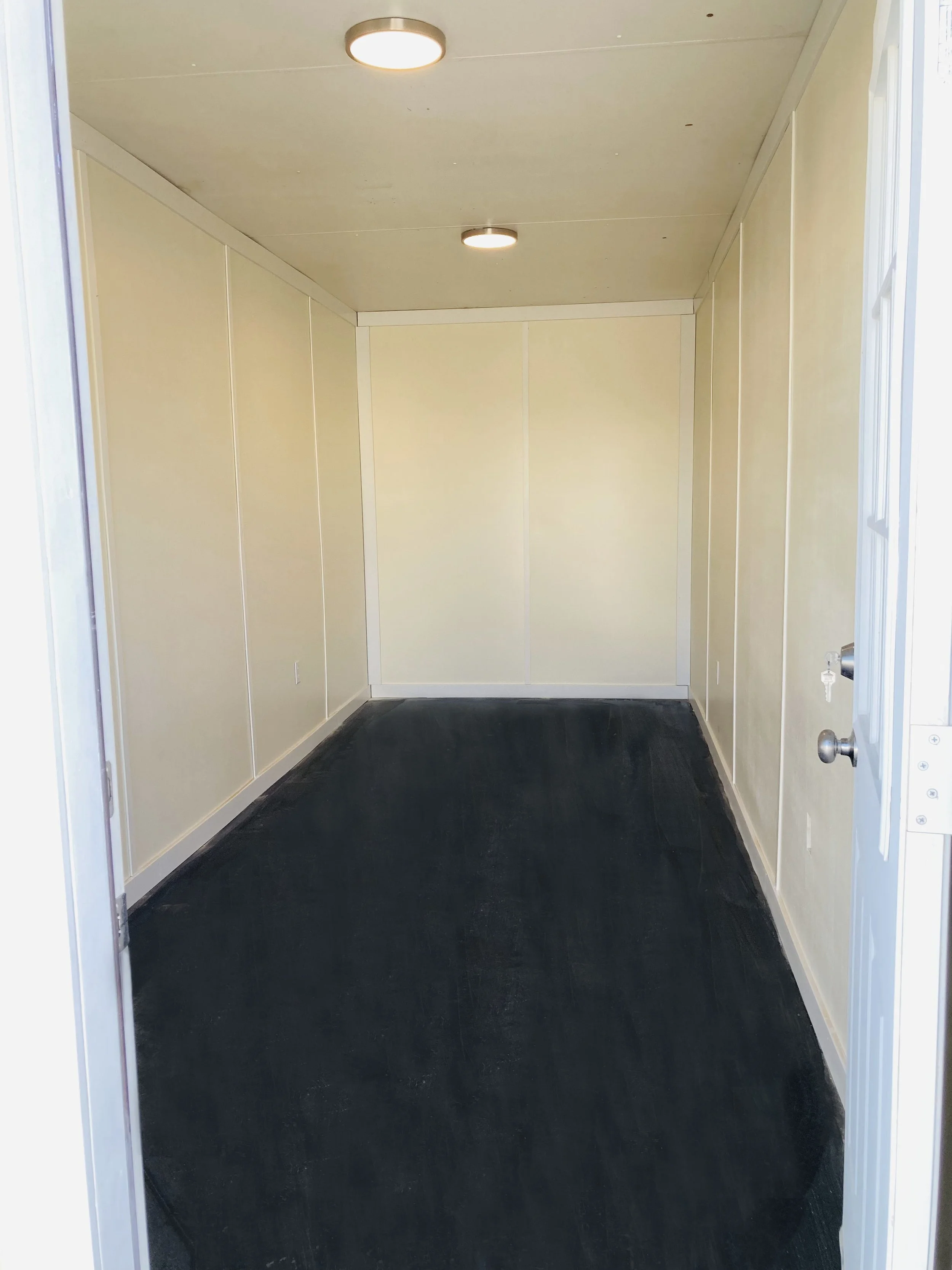 Empty small room with beige walls, black carpet, and ceiling lights, viewed from an open doorway.