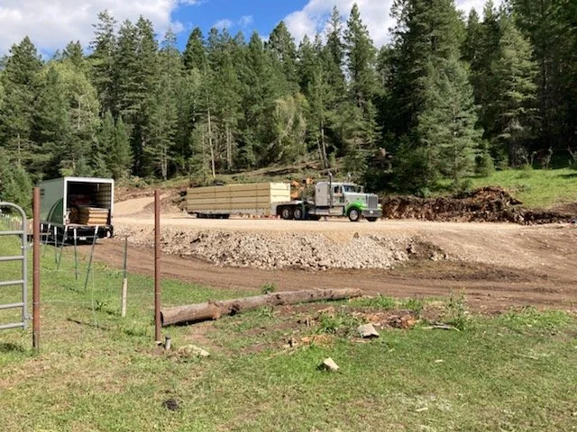 A gray flatbed truck with a green bumper carrying wooden planks on a dirt road, with an open truck trailer nearby, surrounded by green trees and grass.
