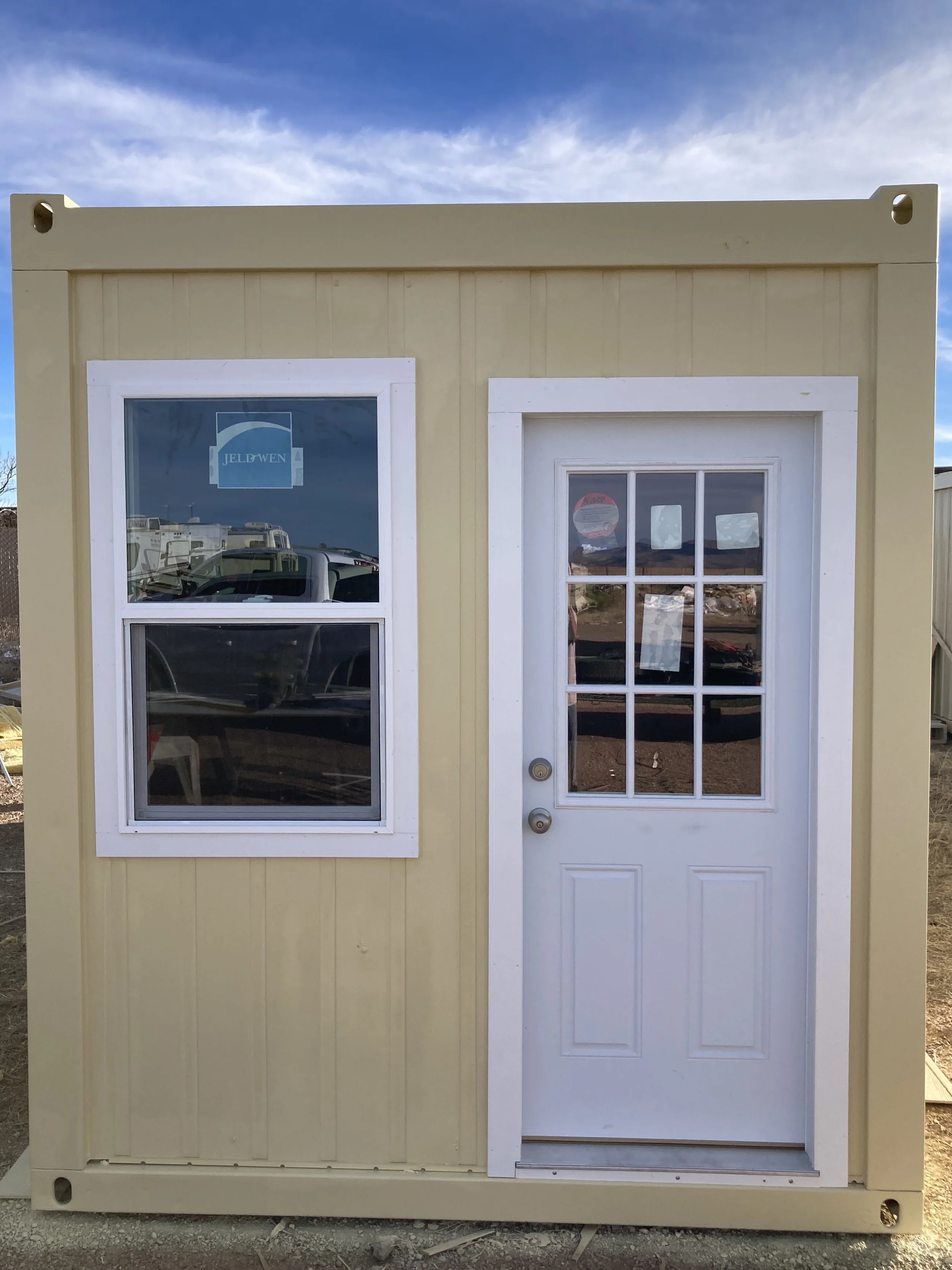 Small beige building with a white door and a window, outdoors with a partly cloudy sky.