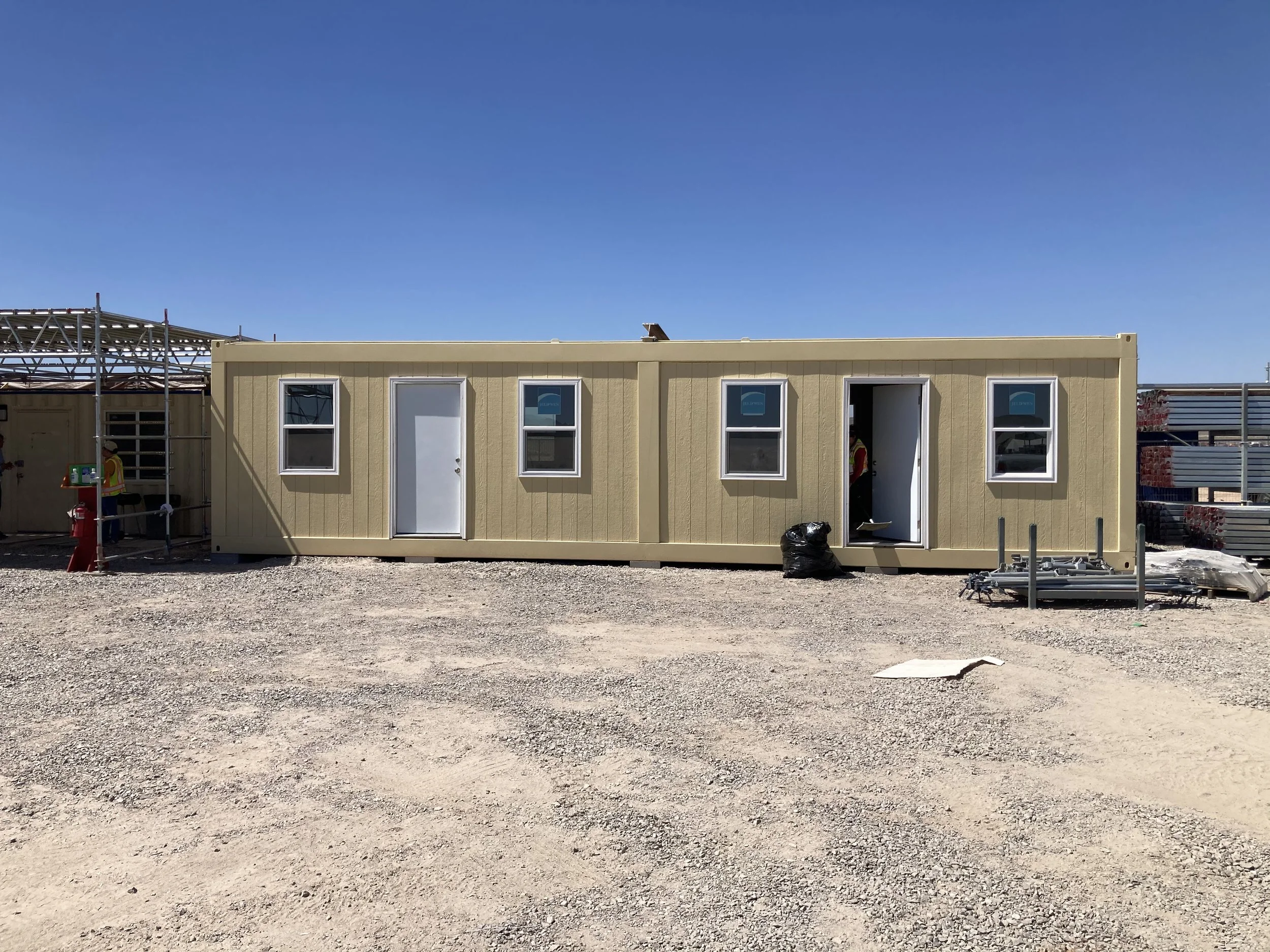 A beige modular building under construction on a gravel lot, with four windows and two doors, some construction materials and workers nearby, under a clear blue sky.