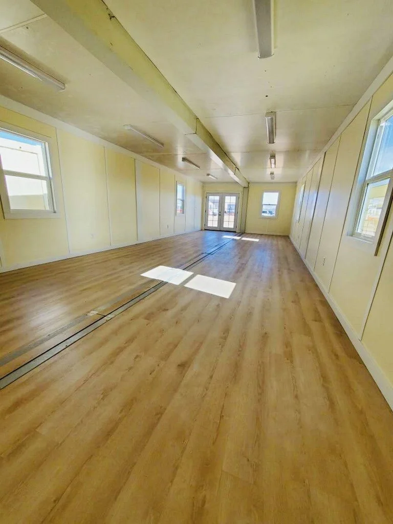 Empty room with light-colored walls, wooden flooring, multiple windows, and a door at the end.