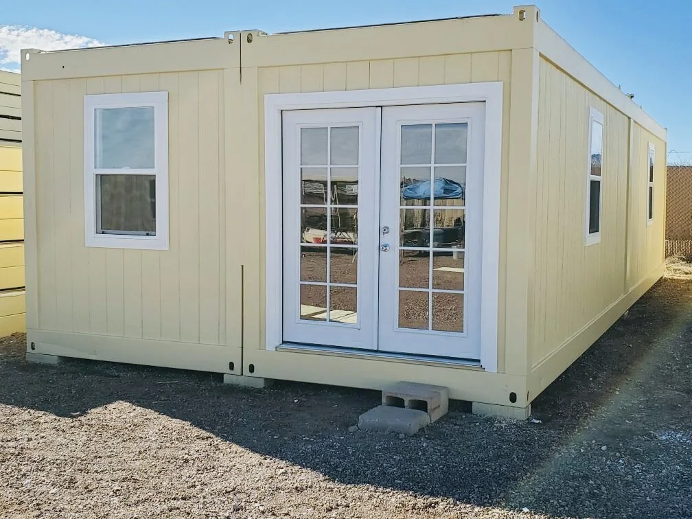 A small, yellow prefabricated building with white-framed windows and French doors, placed on gravel ground with a concrete block at the entrance.