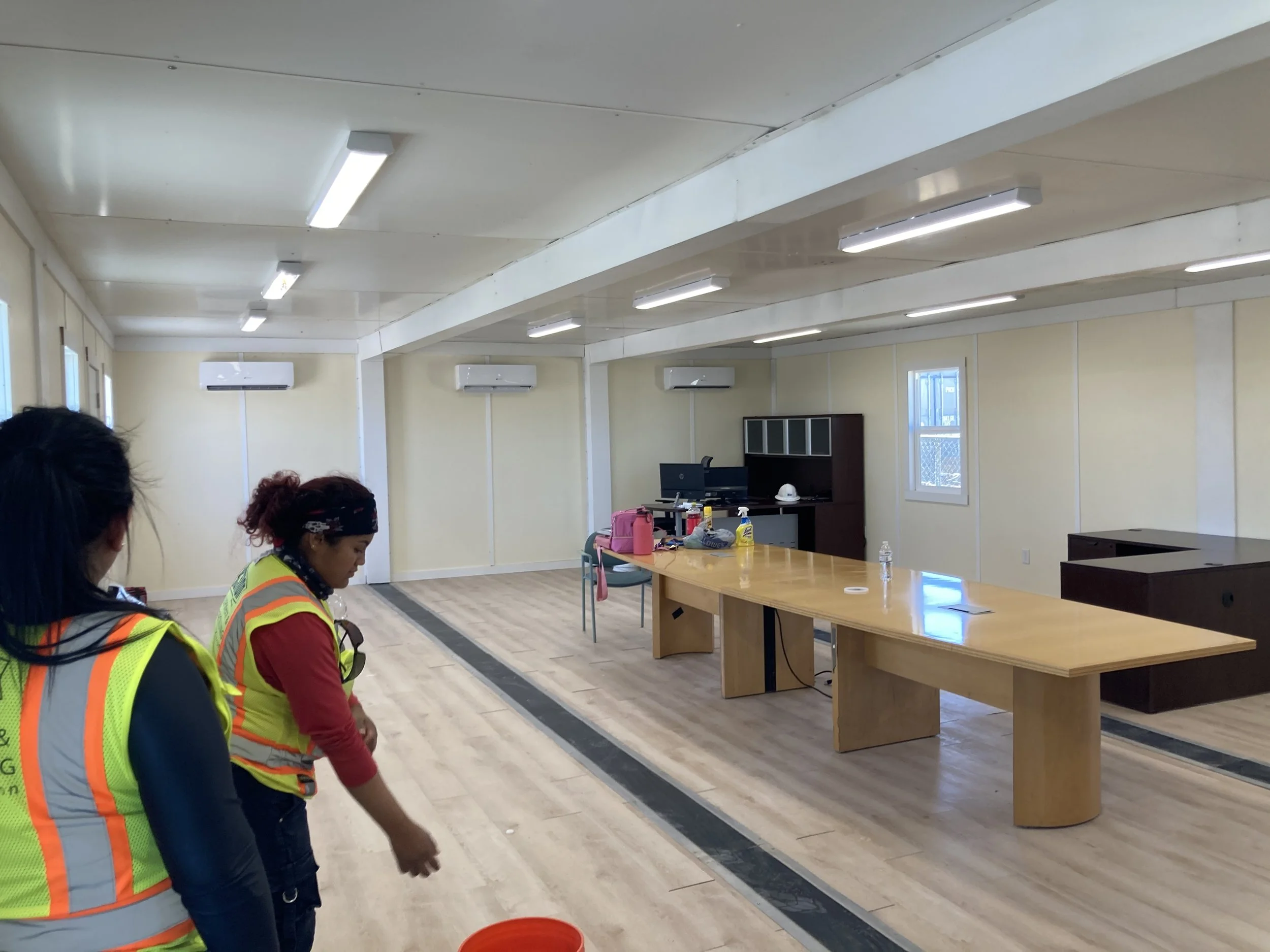 Two women wearing safety vests examining the unfinished interior of a room with a long wooden table and office supplies on it, in a building under construction or renovation.