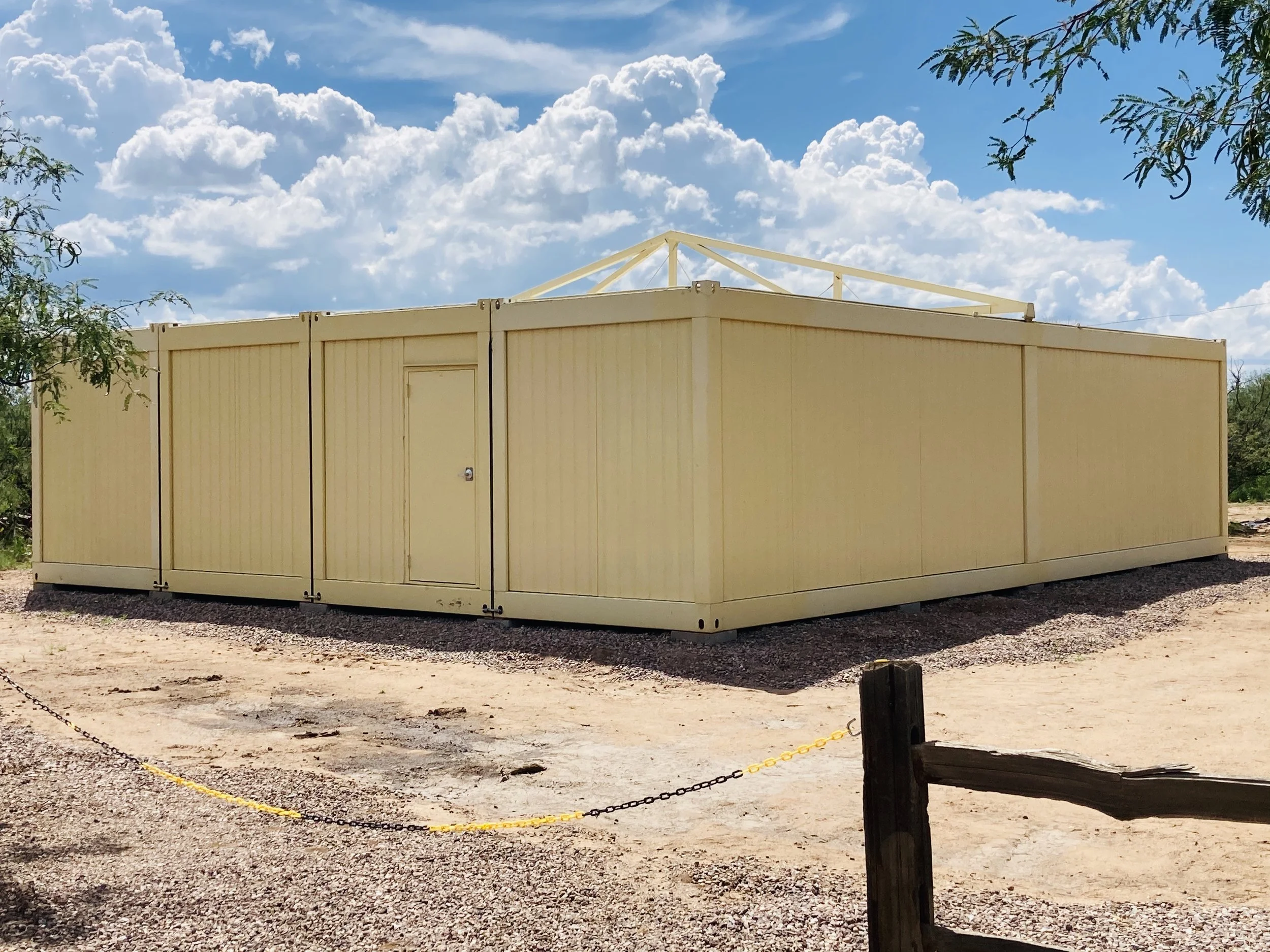 A beige, metal modular building structure with a door, situated outdoors on a gravel ground, under a partly cloudy sky with trees nearby.