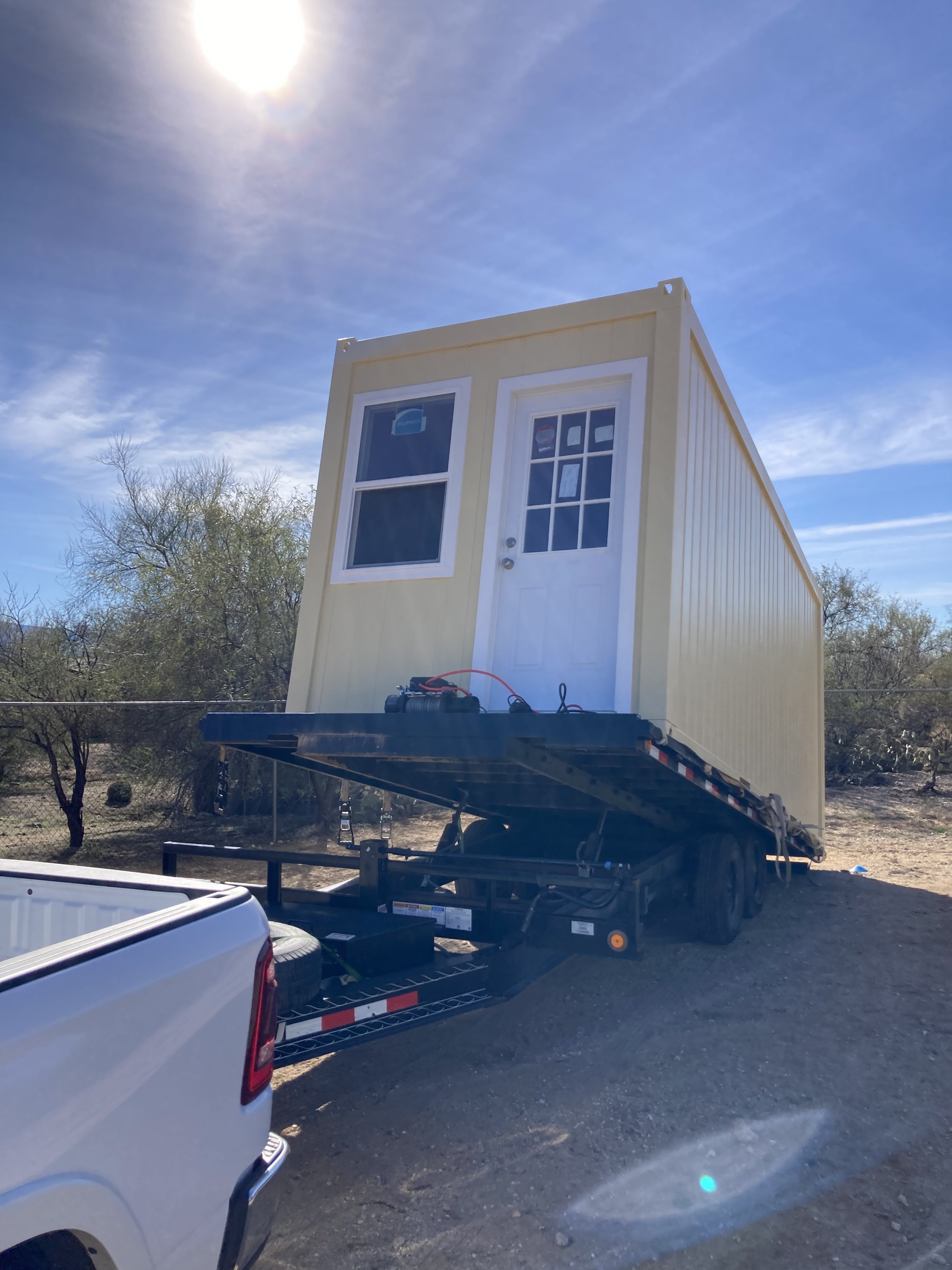 A tiny beige house on a flatbed trailer, attached to a white truck, outdoors with a blue sky and sunlight overhead.