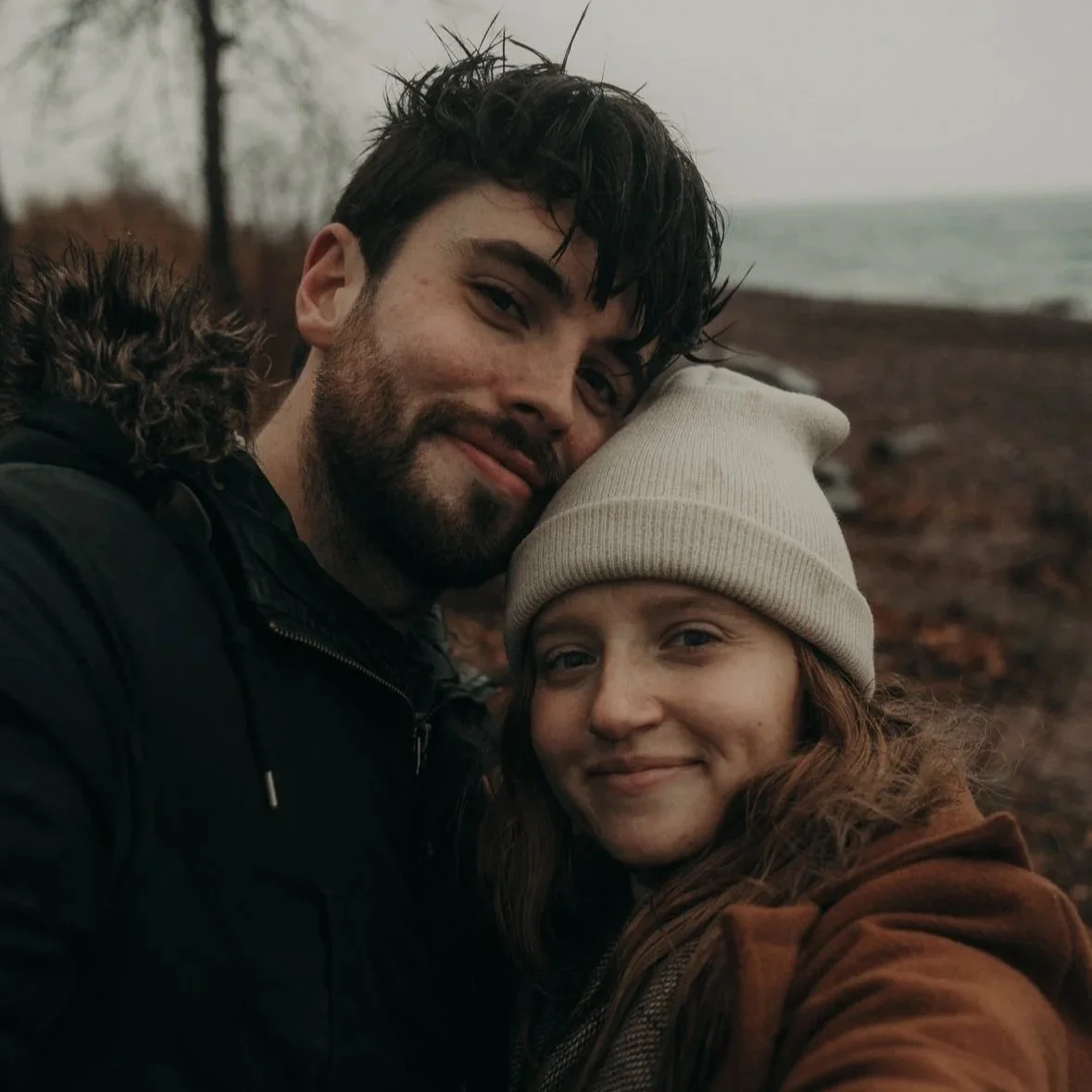 A smiling man and woman taking a selfie outdoors on a cloudy day, with the ocean in the background.