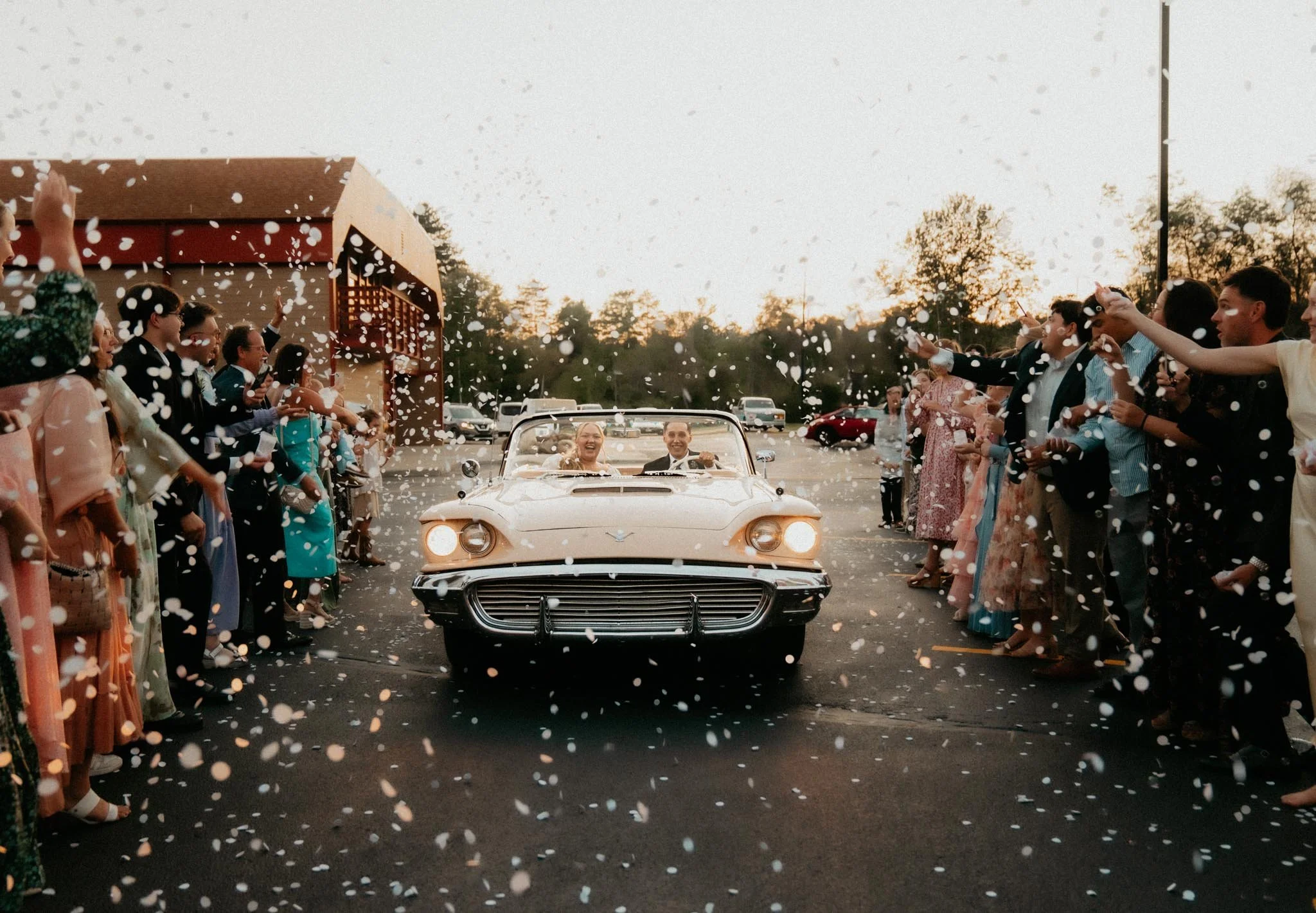 Happy bride and groom driving a white vintage convertible car through a crowd of guests throwing confetti at sunset.