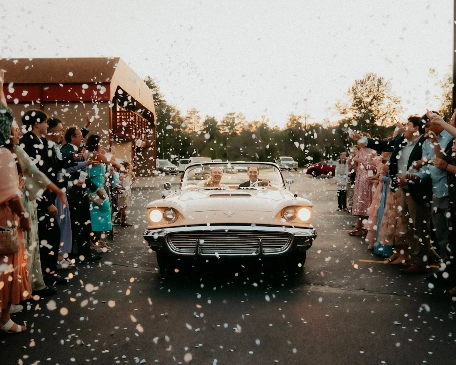 A bride and groom riding in a vintage convertible car through a crowd of well-dressed guests throwing confetti at sunset.
