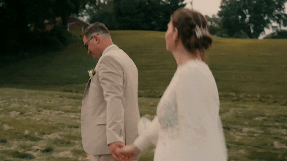 A wedding video of a Minnesota bride and groom holding hands and walking outdoors in a grassy area, dressed in wedding attire, with trees and a hill in the background.