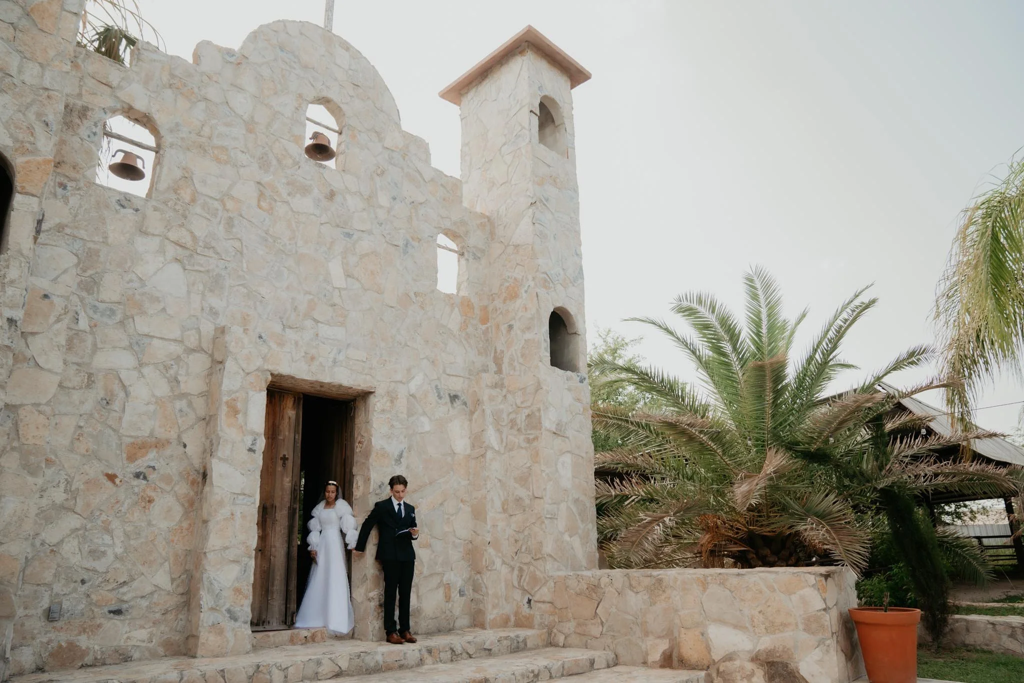 A bride and groom holding hands exiting a stone church with palm trees nearby.