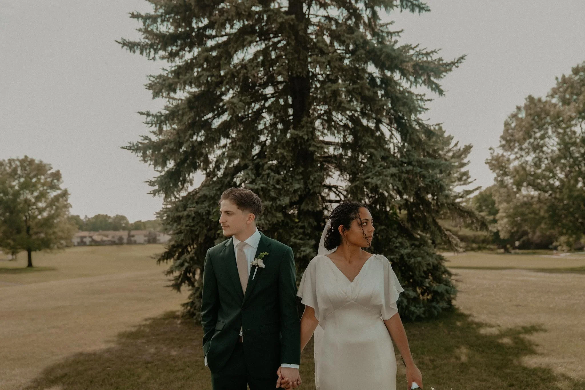 A bride and groom hold hands and stand side by side outdoors, with large evergreen trees in the background during a wedding.