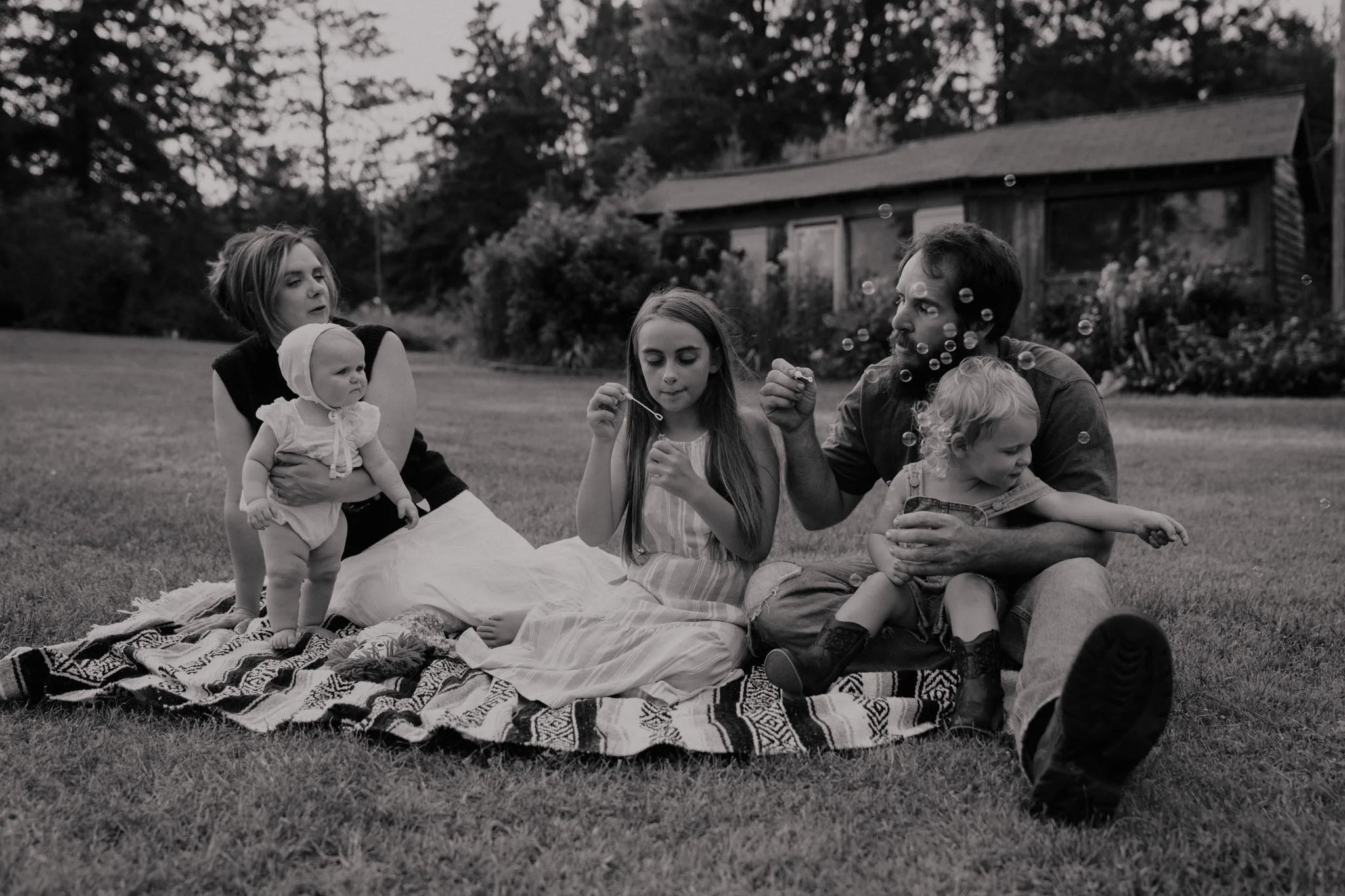 A family sitting on a blanket in a backyard, playing with bubbles on a grassy area with trees and a house in the background.