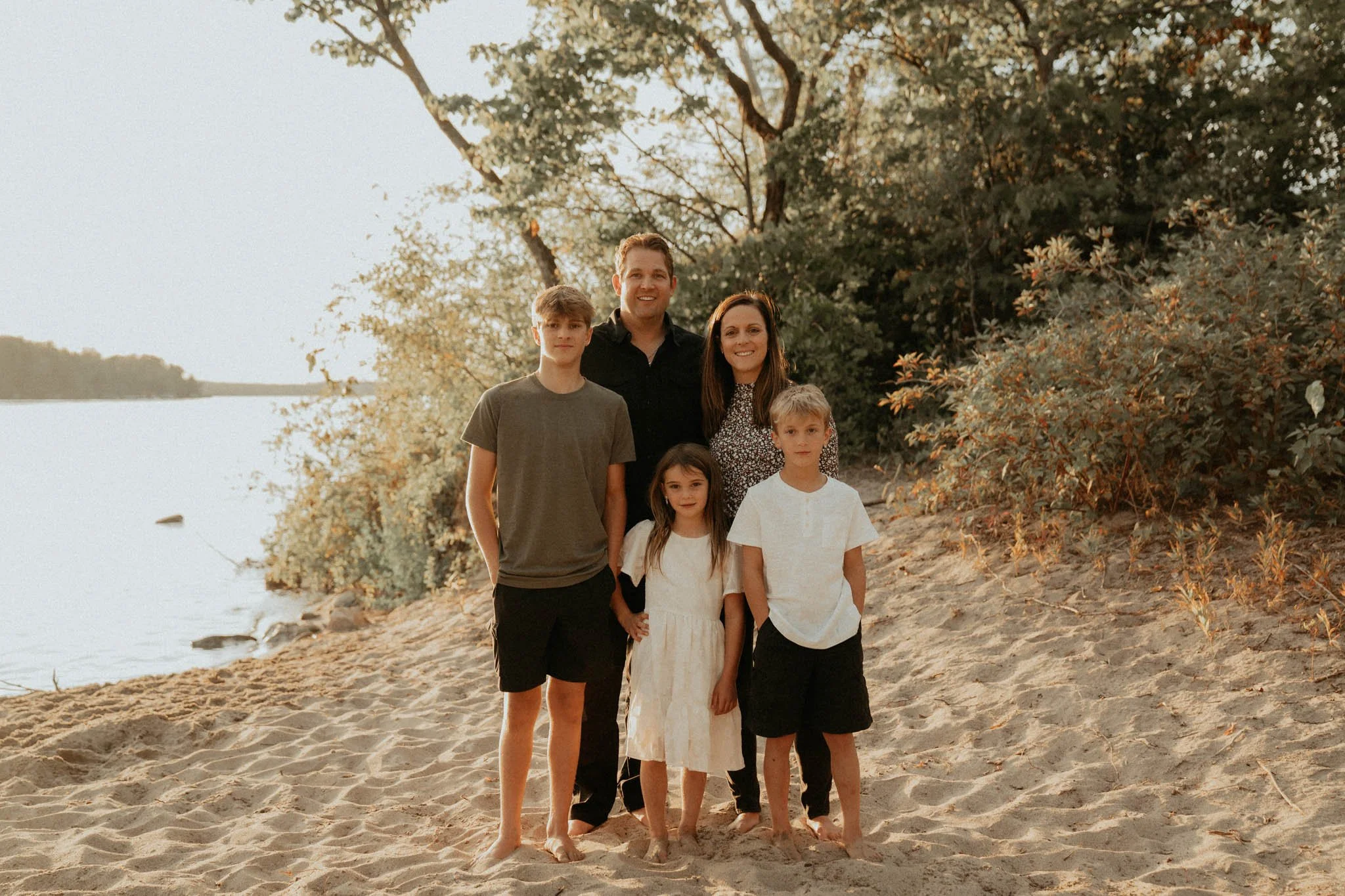 A family of six standing on a sandy beach near a body of water with trees in the background during sunset.