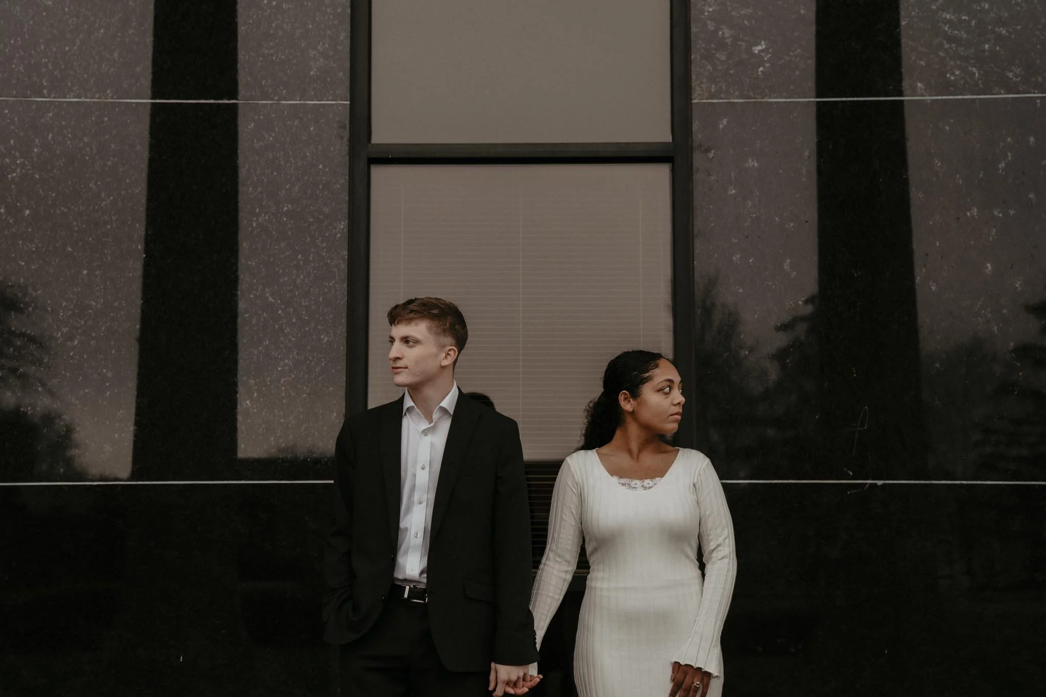 A young man and woman holding hands and standing outside a dark building at night. The man is dressed in a suit and the woman in a white dress, both looking in opposite directions.