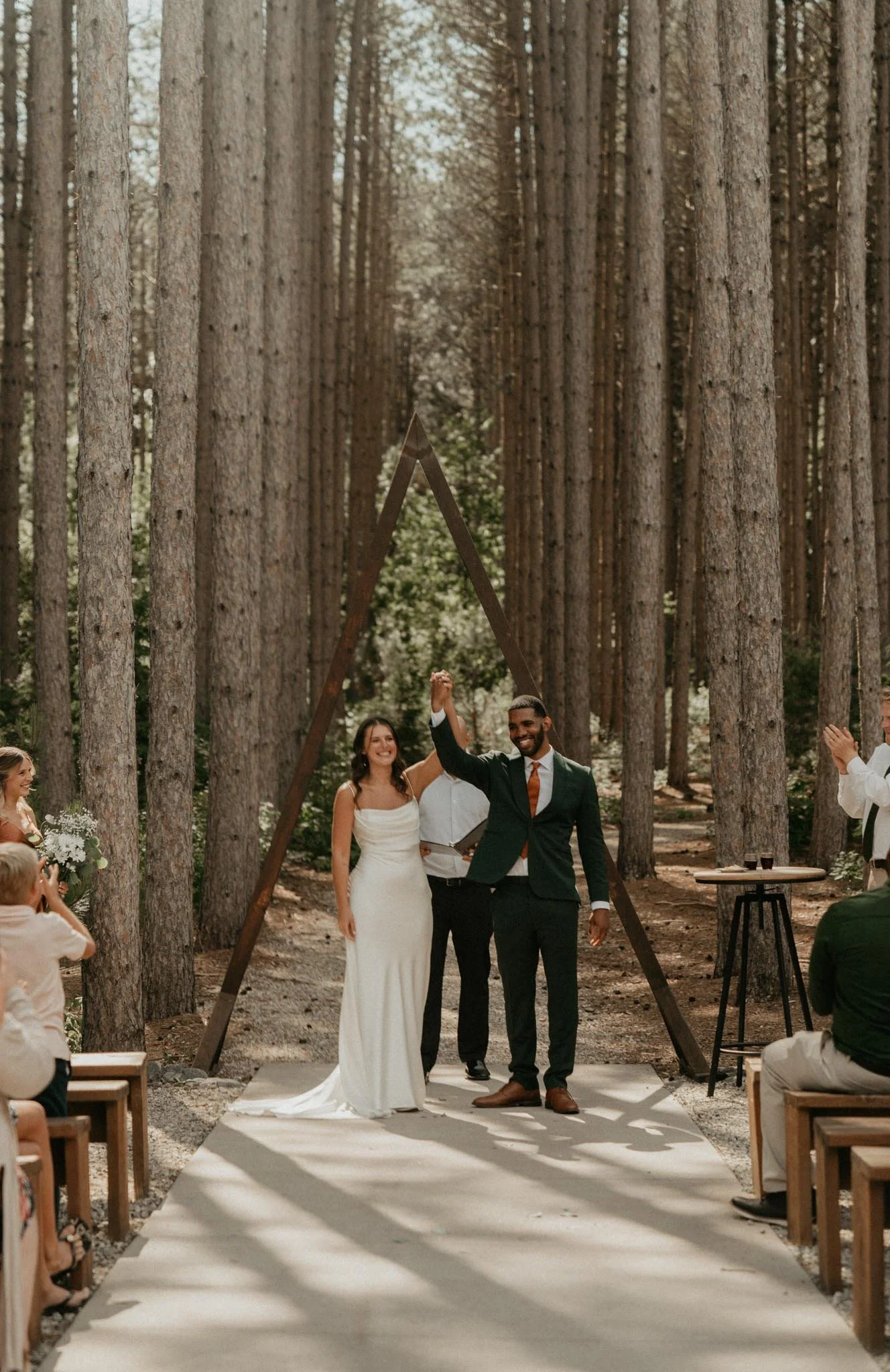 A wedding ceremony taking place outdoors in a forest with tall trees. The bride and groom are standing at the altar, holding hands, smiling. The bride is wearing a white wedding dress, while the groom is in a dark suit with a orange tie. Guests are s