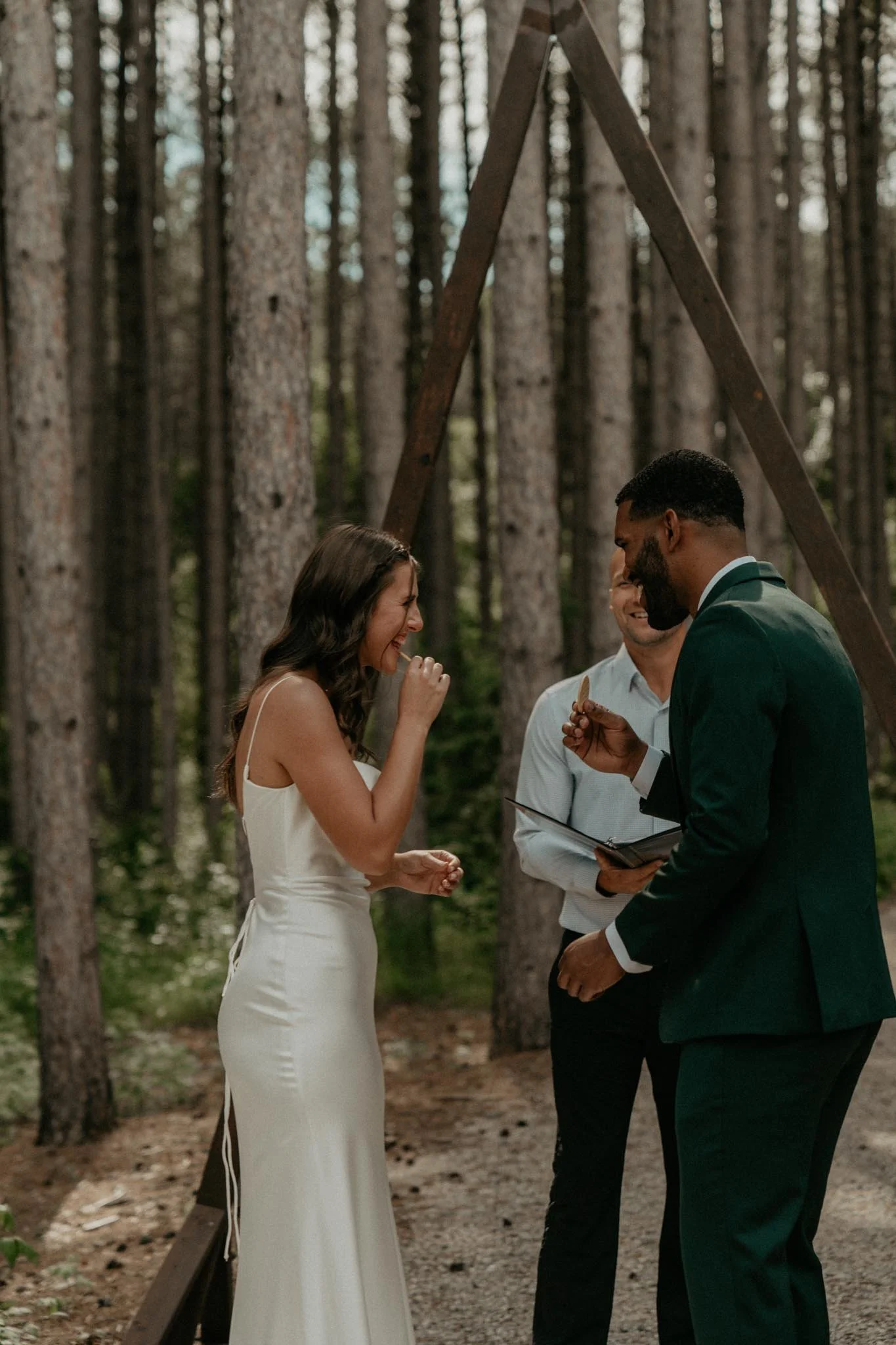 A couple exchange vows during their outdoor wedding ceremony in a forest. A woman in a white dress is visibly emotional, and a man in a dark green suit is holding a ring. An officiant stands between them with a book.