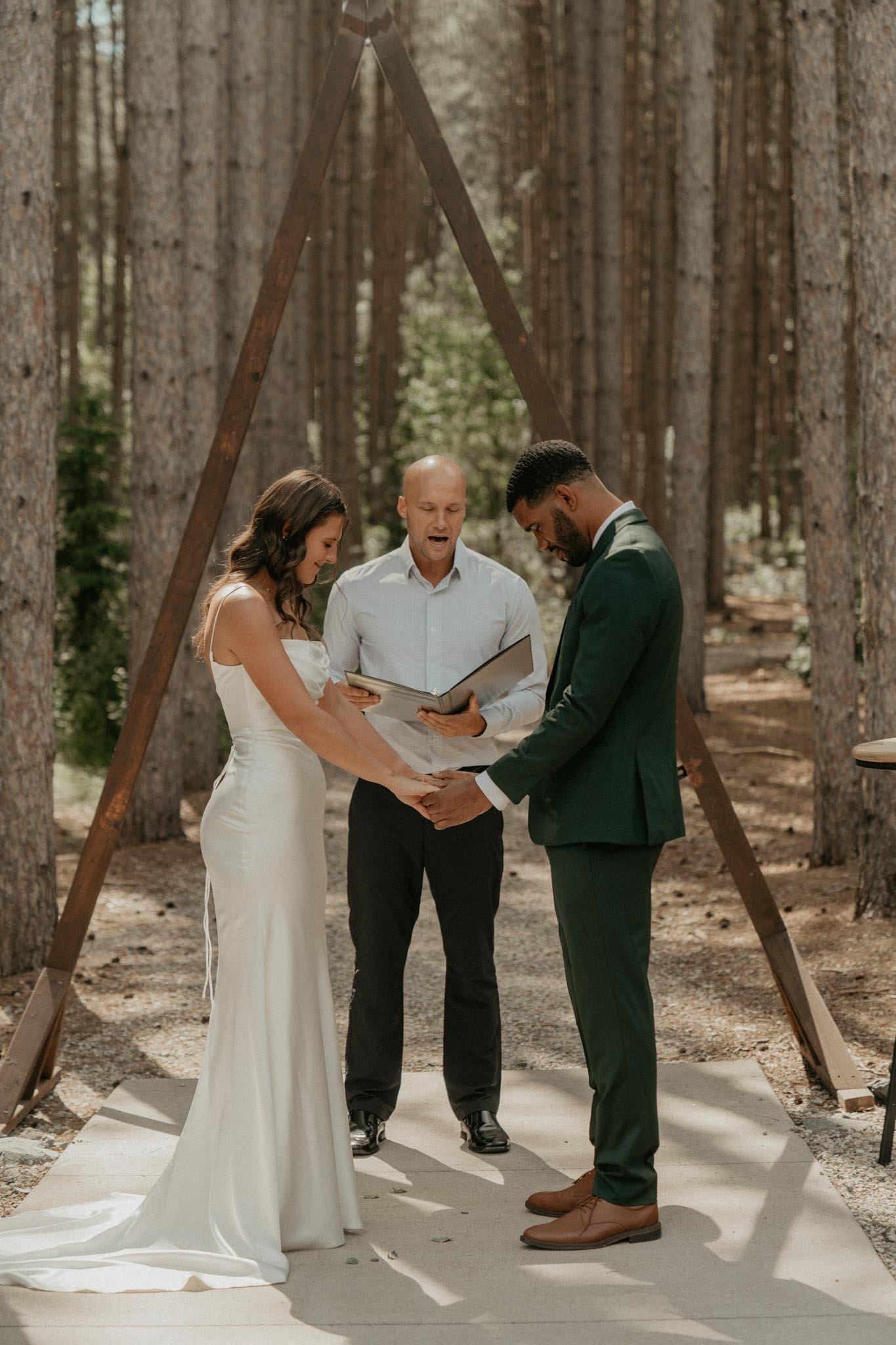 A couple exchanging vows at a forest wedding ceremony. The bride is wearing a white wedding dress, and the groom is in a dark green suit. An officiant stands behind them, holding an open book, as they hold hands under a wooden wedding arch.