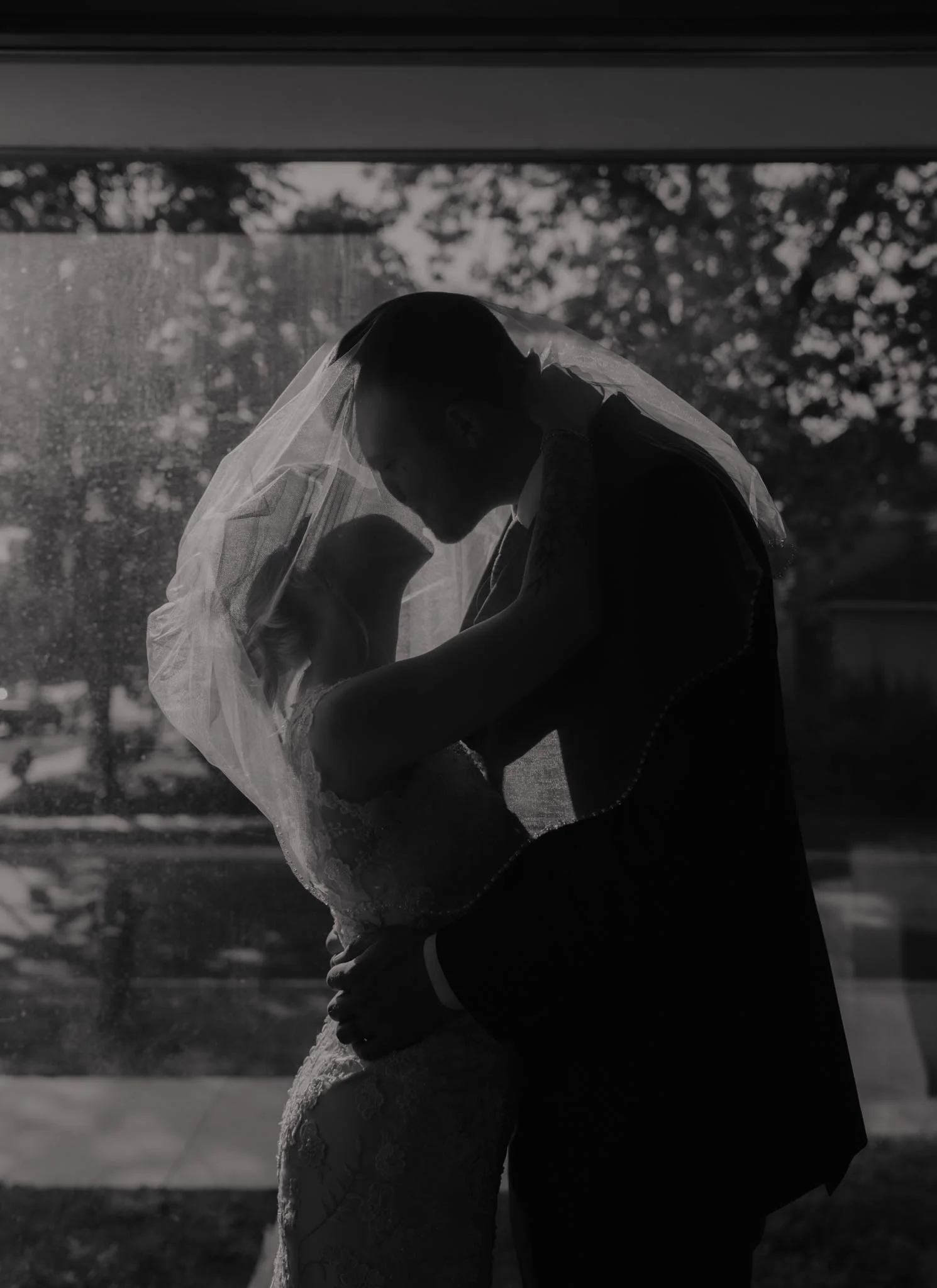 Silhouette of a bride and groom embracing and kissing behind a window, with a veil on the bride's head and wedding attire, outdoors during daytime.