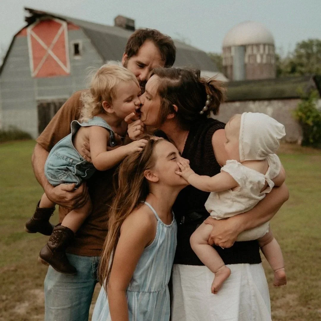 A family of five sharing a close and affectionate moment outdoors, with a barn and silo in the background.