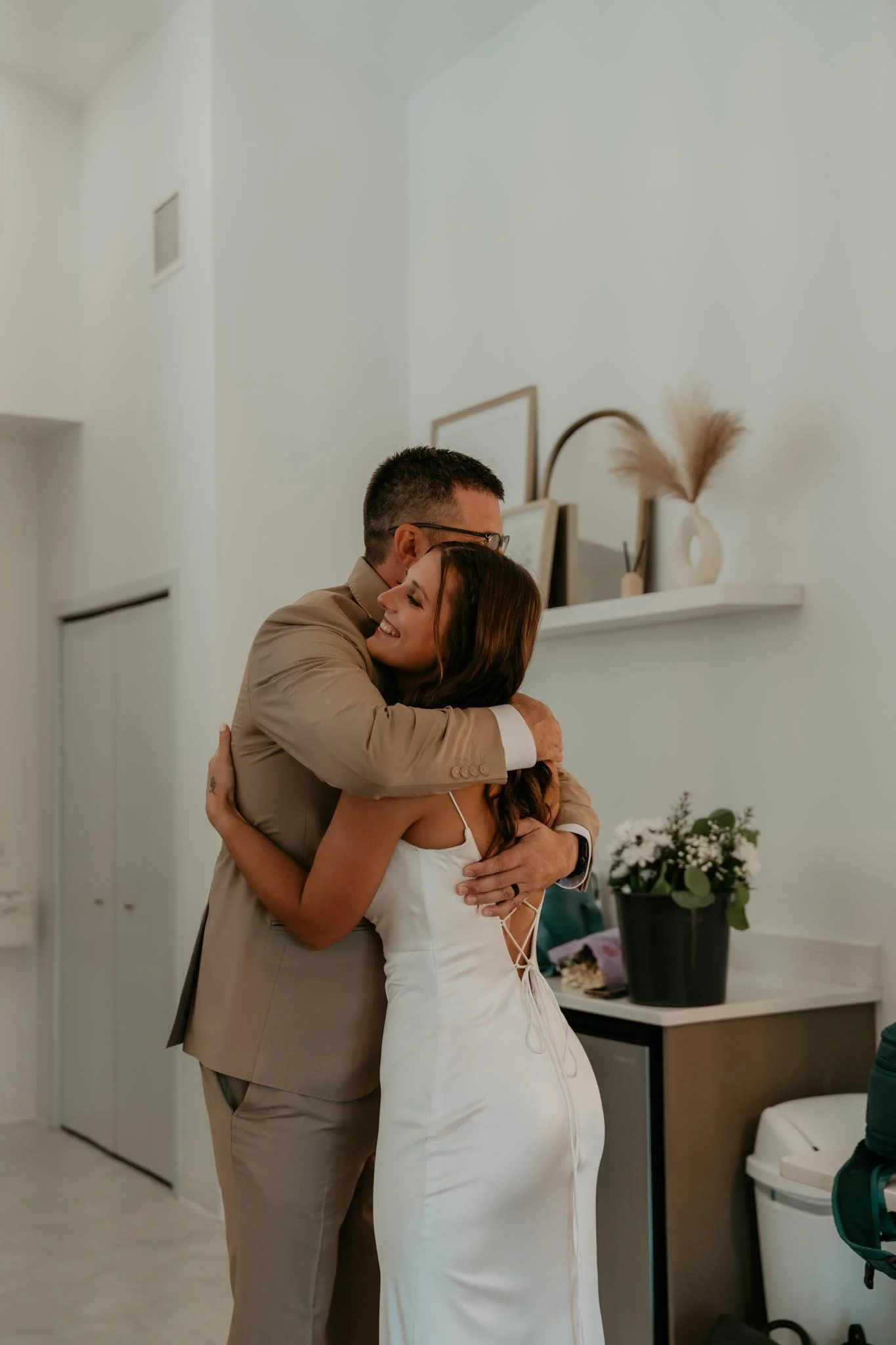 A man and woman hugging each other happily indoors, the woman is wearing a white dress, and the man is dressed in a beige suit.