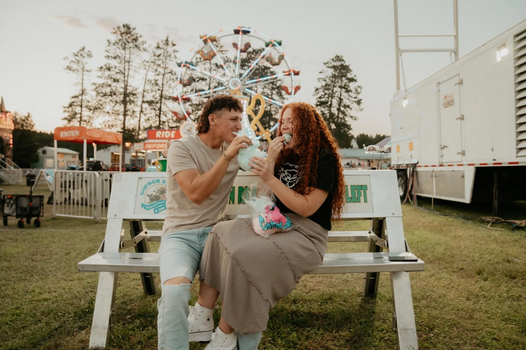 A young man and woman sit on a bench at a fairground, sharing cotton candy and smiling at each other, with a Ferris wheel and amusement rides in the background during sunset.