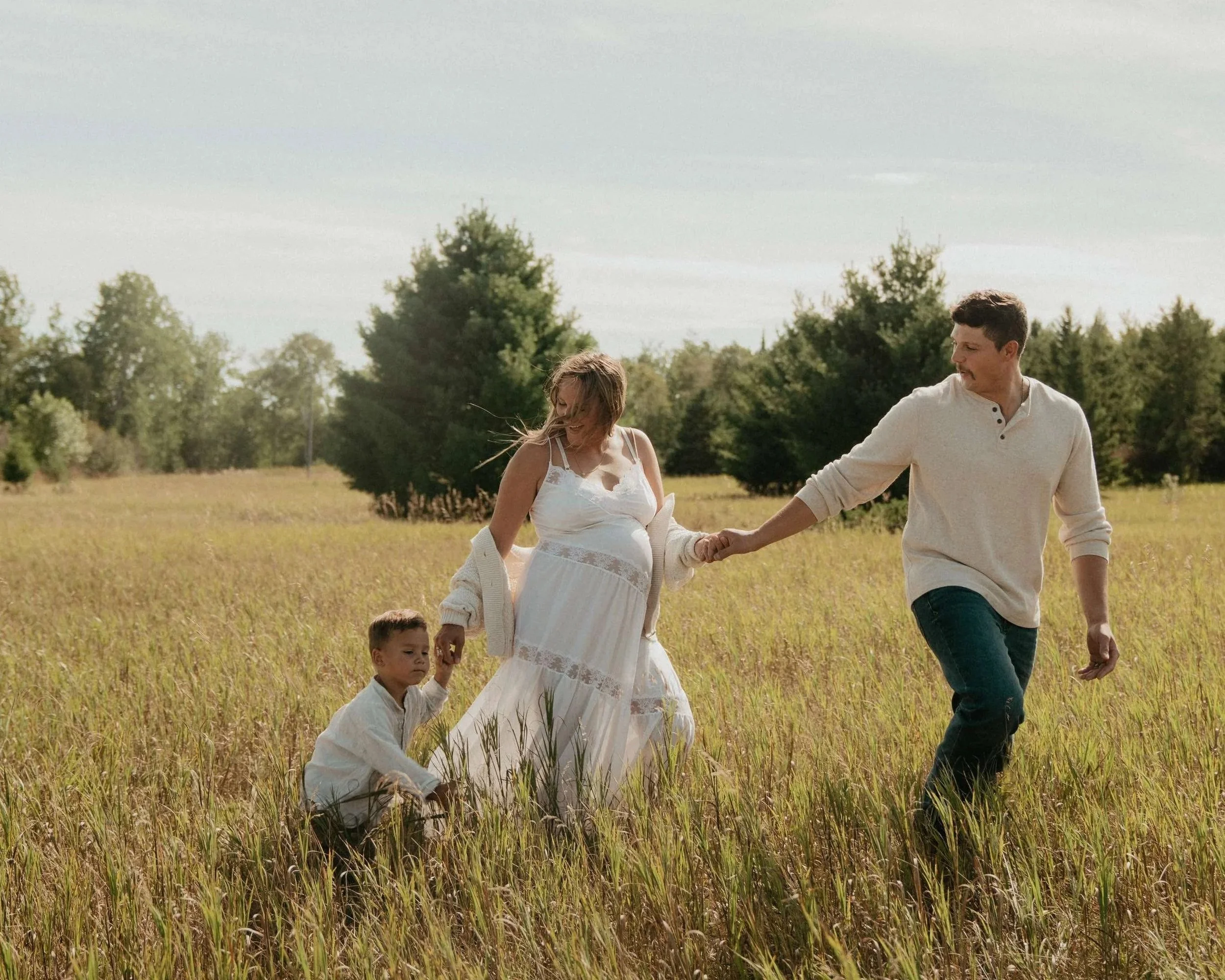 A family of four walking through a grassy field with trees in the background, holding hands and enjoying a sunny day.