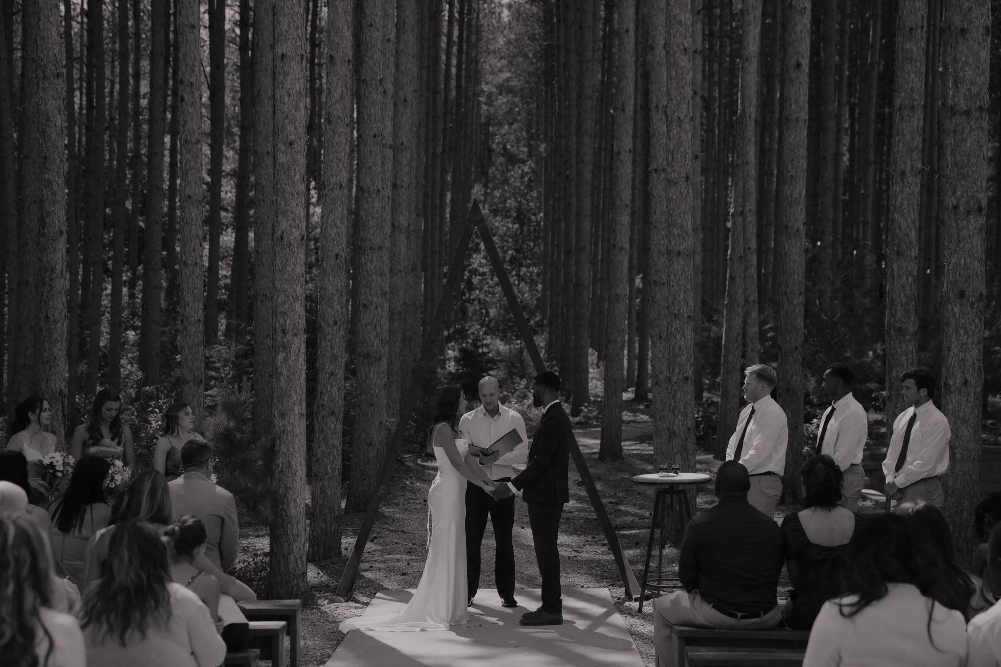 A black and white photo of a wedding in a forest with a couple holding hands and being married by officiant, surrounded by guests, with tall trees in the background.