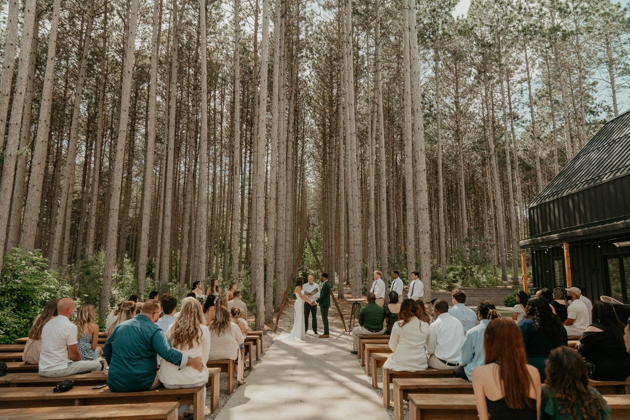 Wedding ceremony with bride and groom exchanging vows under a tall pine forest, with guests seated on wooden benches on both sides of a white aisle.