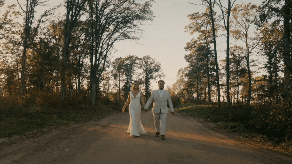 A couple in wedding attire walking hand in hand along a dirt path surrounded by trees at sunset during their wedding videography..