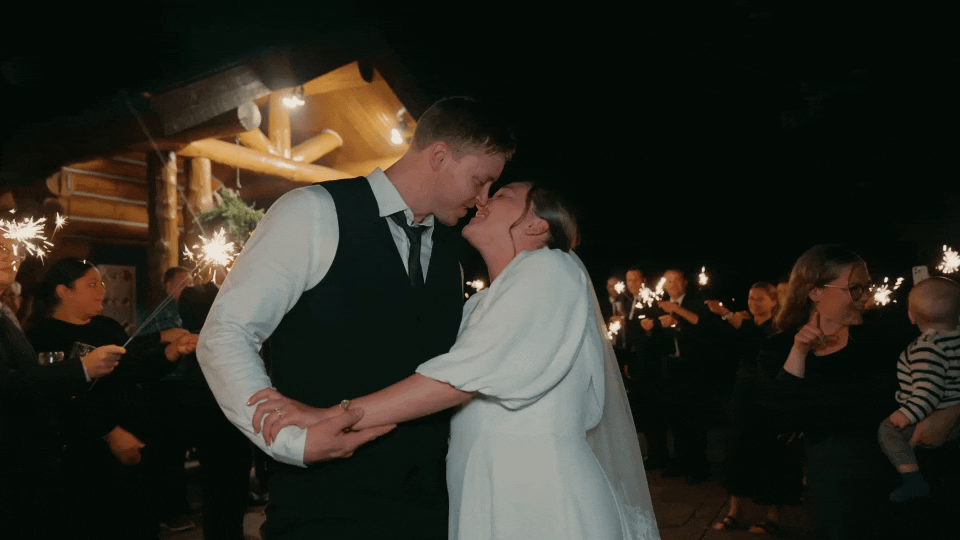 A couple is kissing during a celebration, holding hands, with sparklers held by guests in the background at night during their wedding videography..