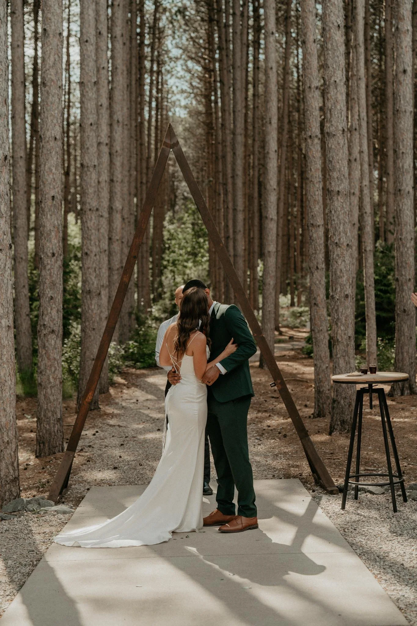 A couple getting married in an outdoor forest setting, under a wooden triangular arch, with an officiant standing nearby