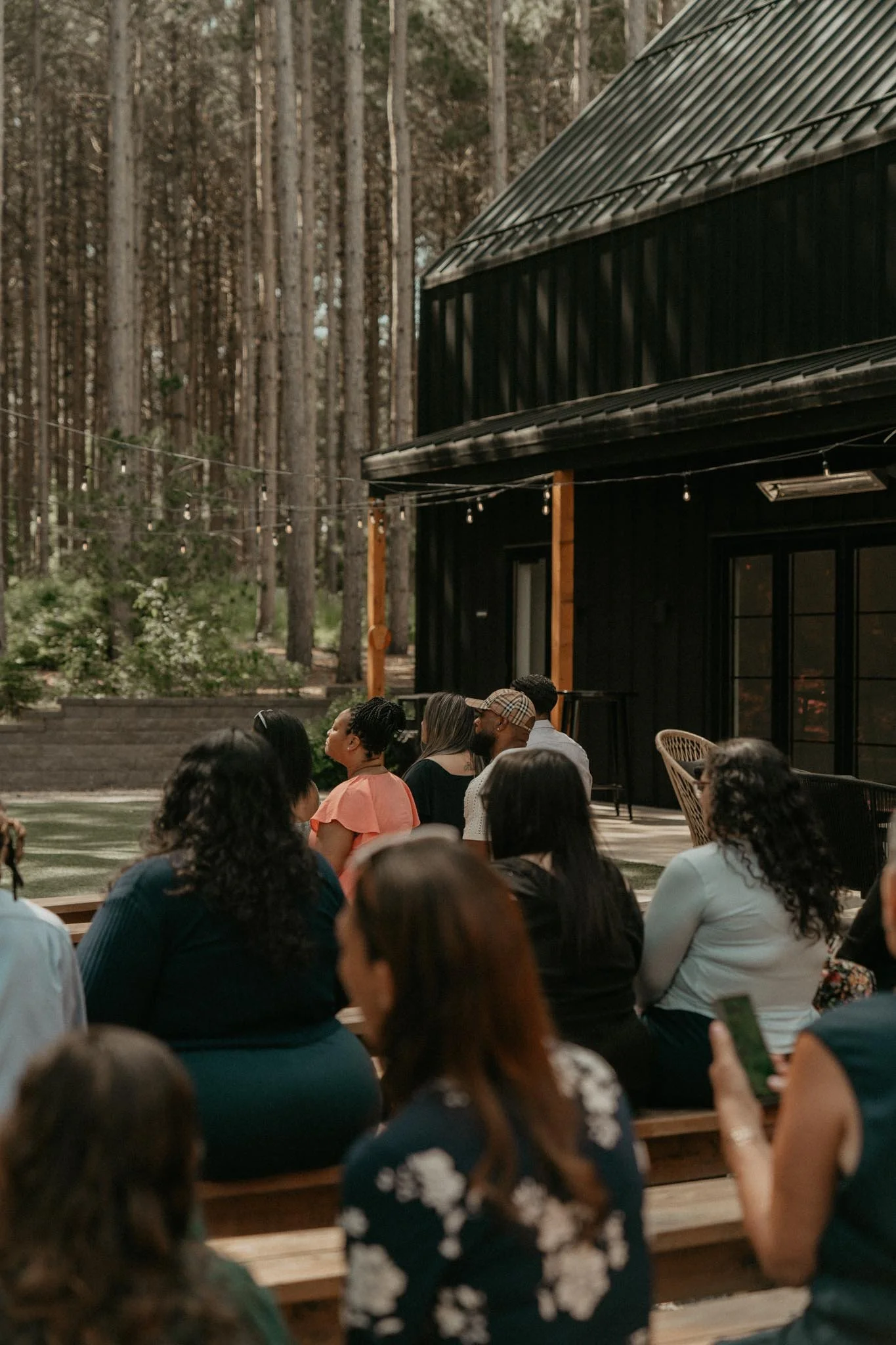 People sitting outdoors in front of a black building with string lights and trees in the background.
