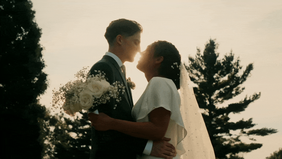 An Indiana wedding videography couple in wedding attire sharing a kiss outdoors during sunset, with trees in the background.