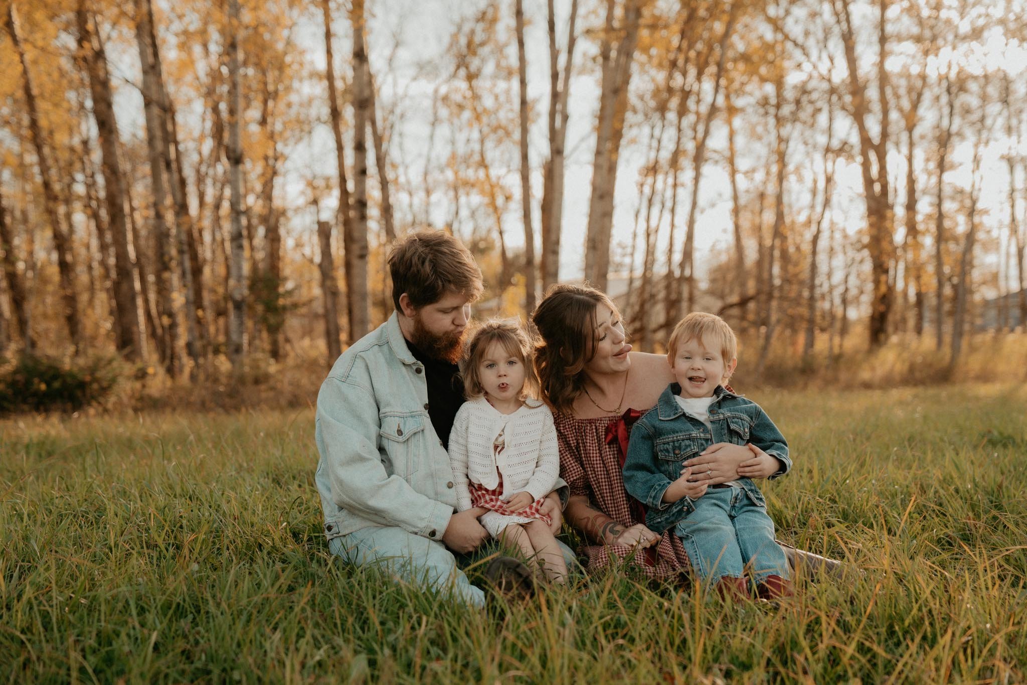 Family of four sitting on grass in a park with autumn trees in the background.