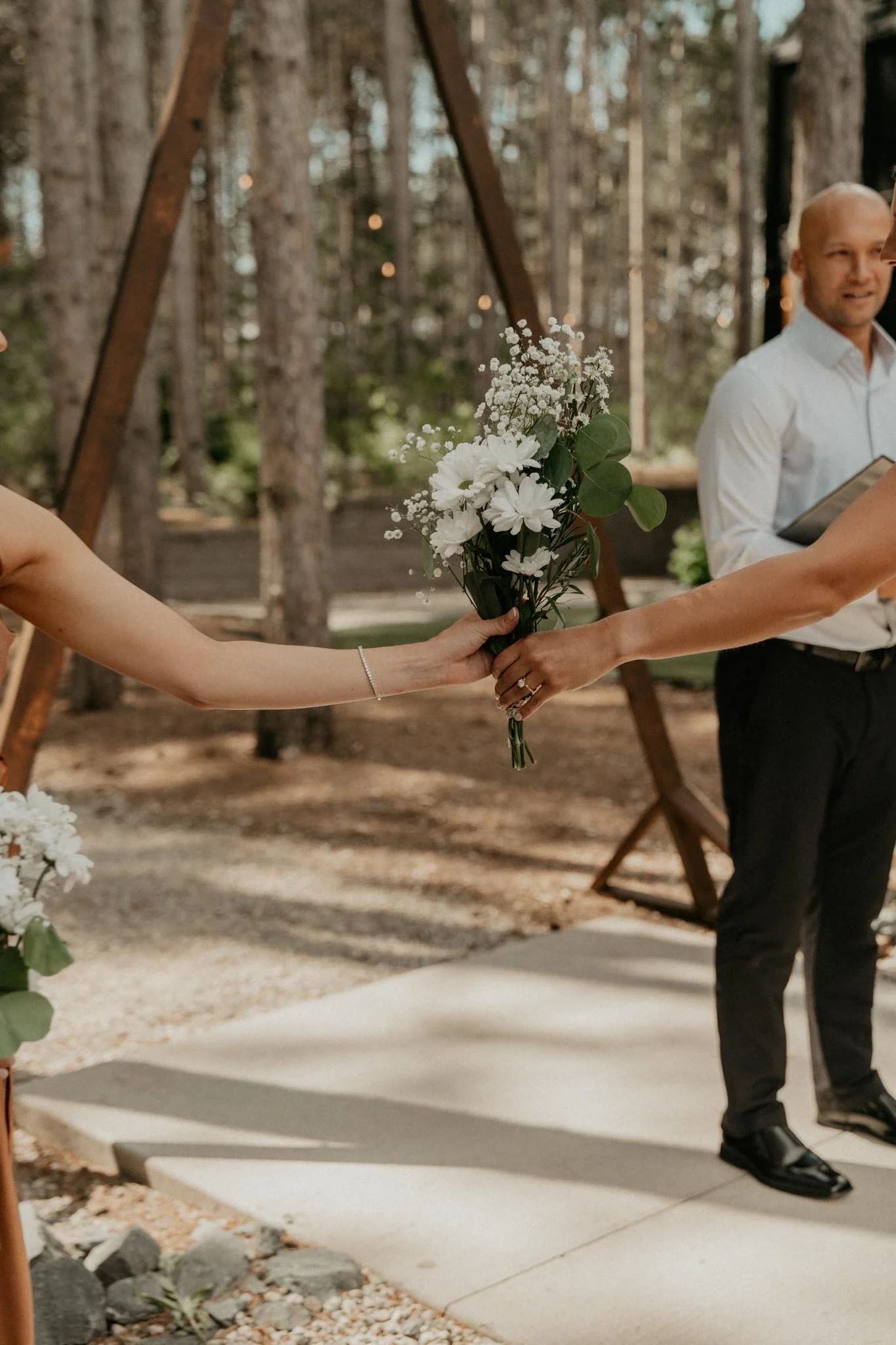 Two individuals exchanging a bouquet of white flowers during a wedding ceremony outdoors in a wooded area.