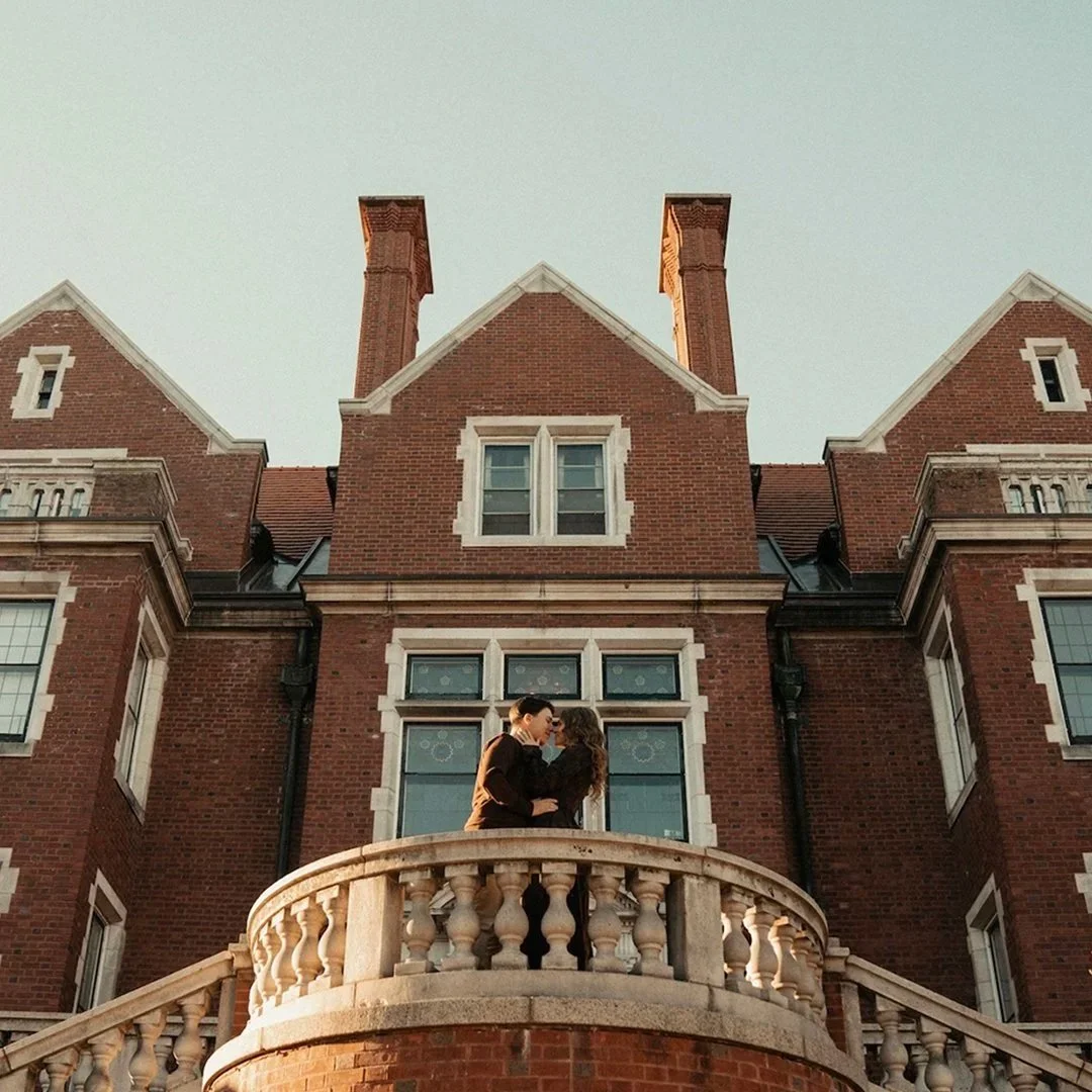 A romantic couple embracing on a balcony in front of an ornate, historic brick mansion.
