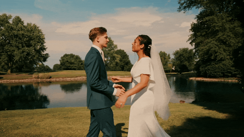 A wedding video shot of a bride and groom are holding hands and facing each other outdoors near a lake with trees in the background during their wedding ceremony.