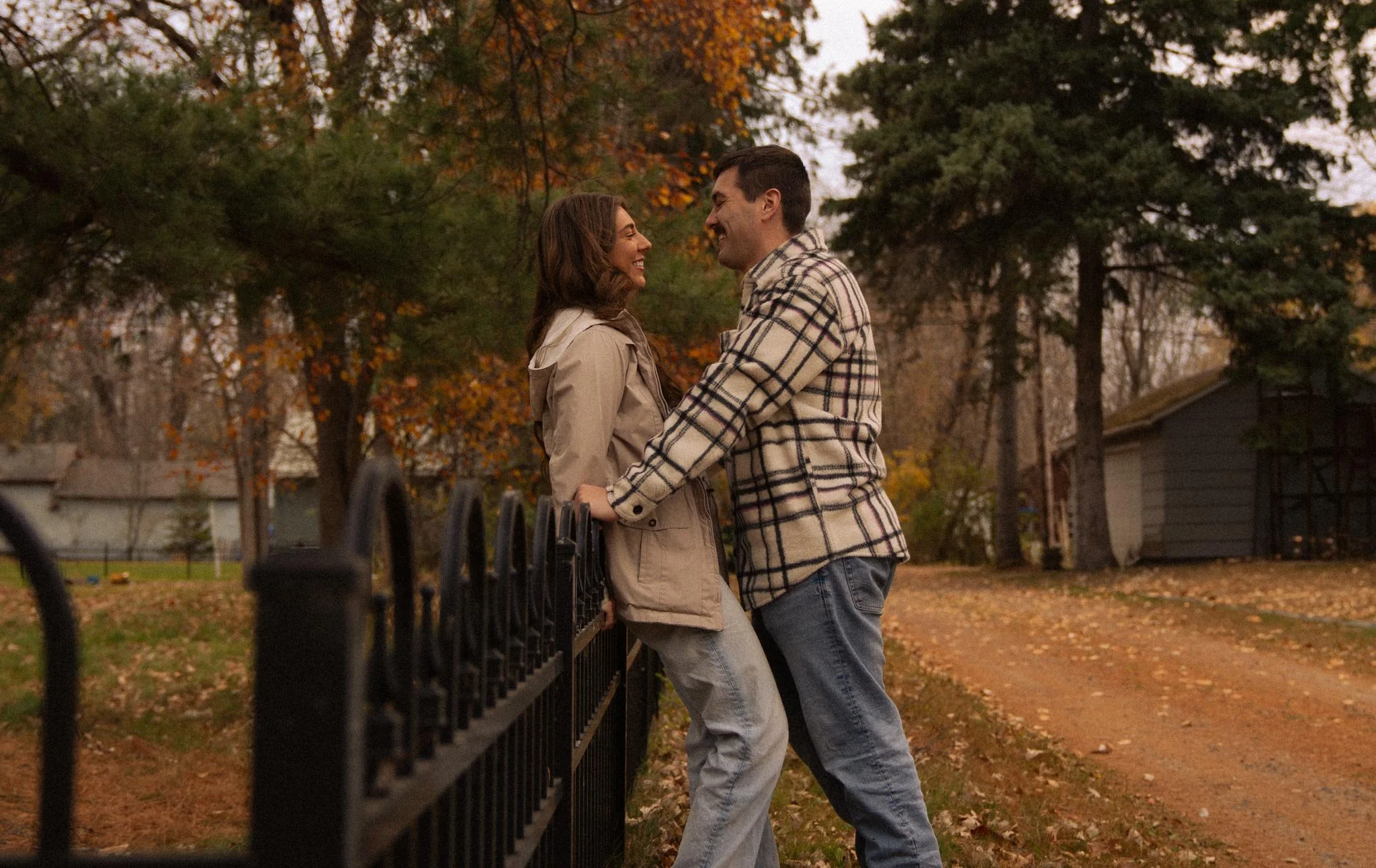 A couple standing close and smiling at each other outdoors during autumn, with trees and houses in the background.