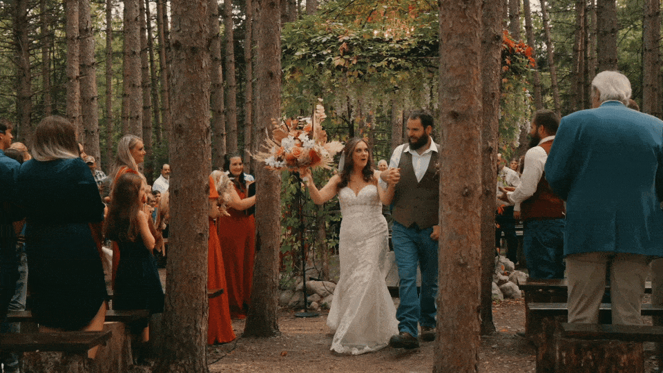 Bride and groom walking down the aisle during their wedding in a forested outdoor wedding ceremony, surrounded by guests.