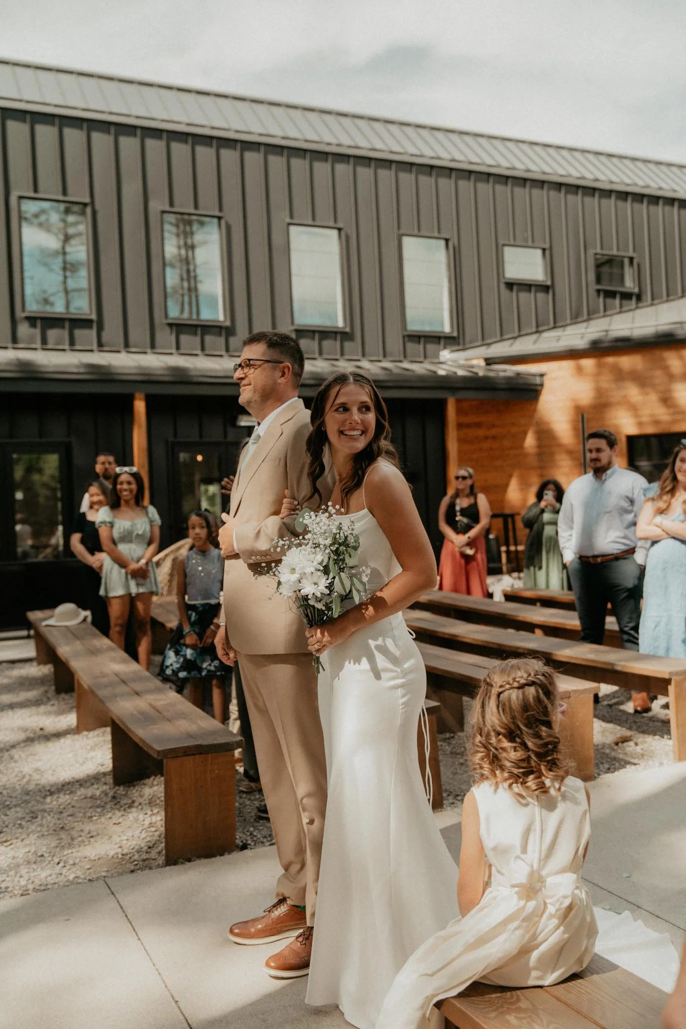 Bride in a white dress holding a bouquet, standing next to a man in a tan suit, at an outdoor wedding ceremony. Guests seated on benches in the background.