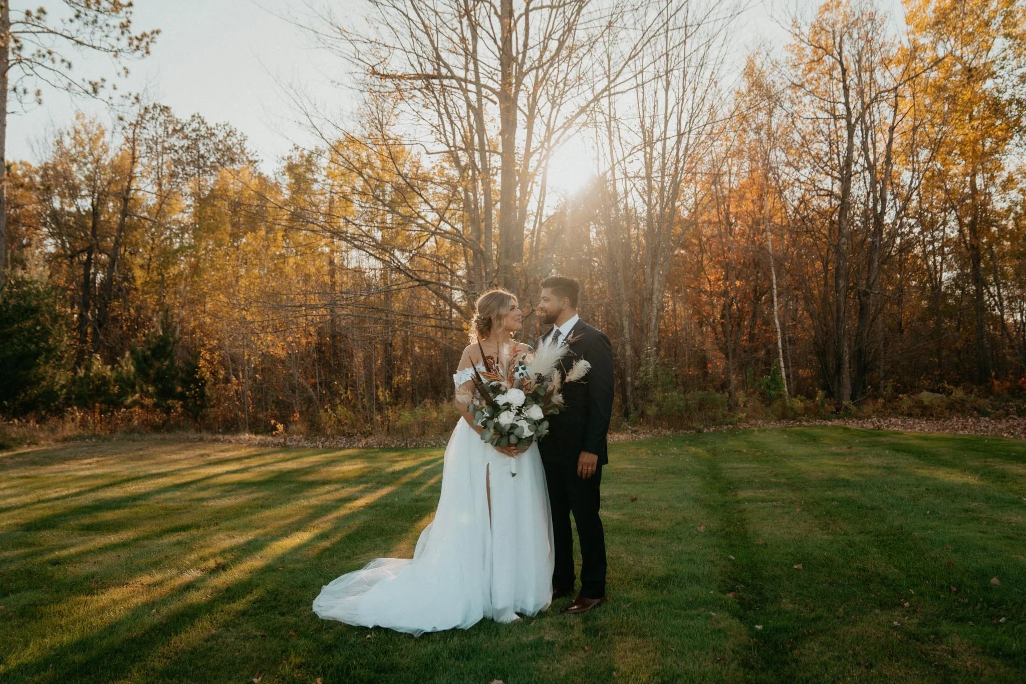 A bride and groom standing on a grassy field at sunset with trees in fall foliage in the background, holding a bouquet and looking at each other.
