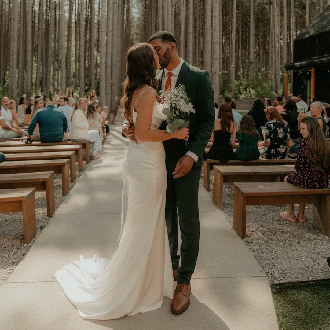 A bride and groom kiss at their outdoor wedding ceremony in a wooded area, with guests seated on wooden benches and trees in the background.